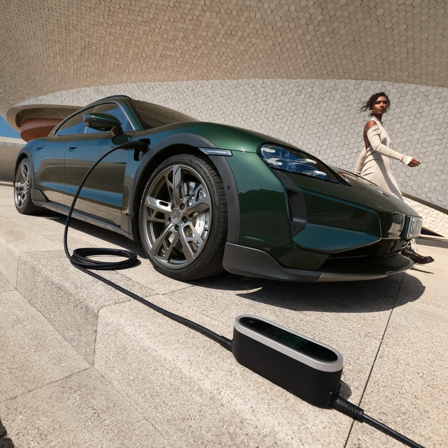 Porsche Taycan vert en charge sur une plateforme en béton, avec une femme en robe beige marchant à proximité.