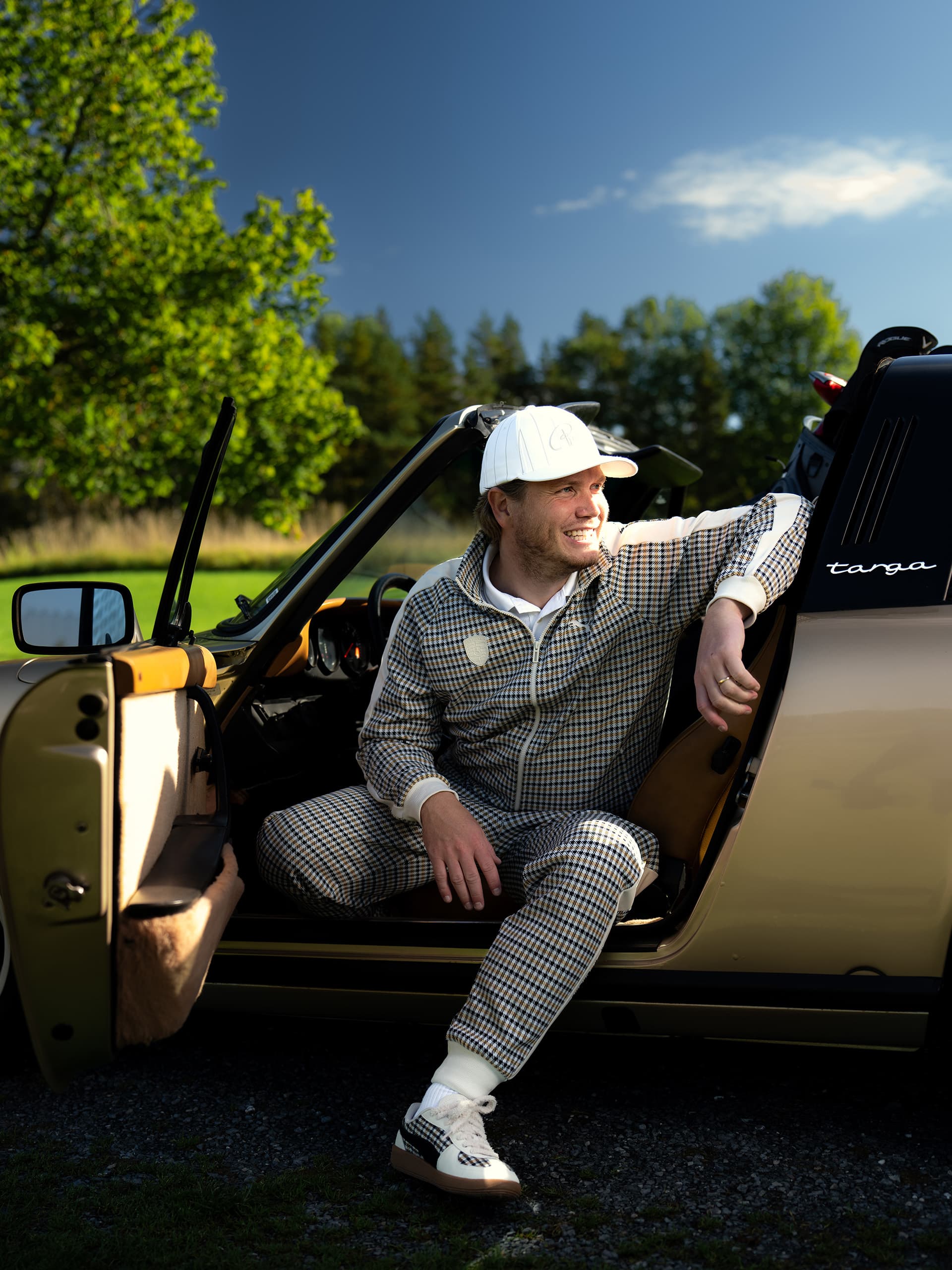 Mann in kariertem Outfit und weißer Mütze sitzt in einem goldenen Porsche Targa, lächelnd unter klarem blauen Himmel mit grünen Bäumen im Hintergrund.