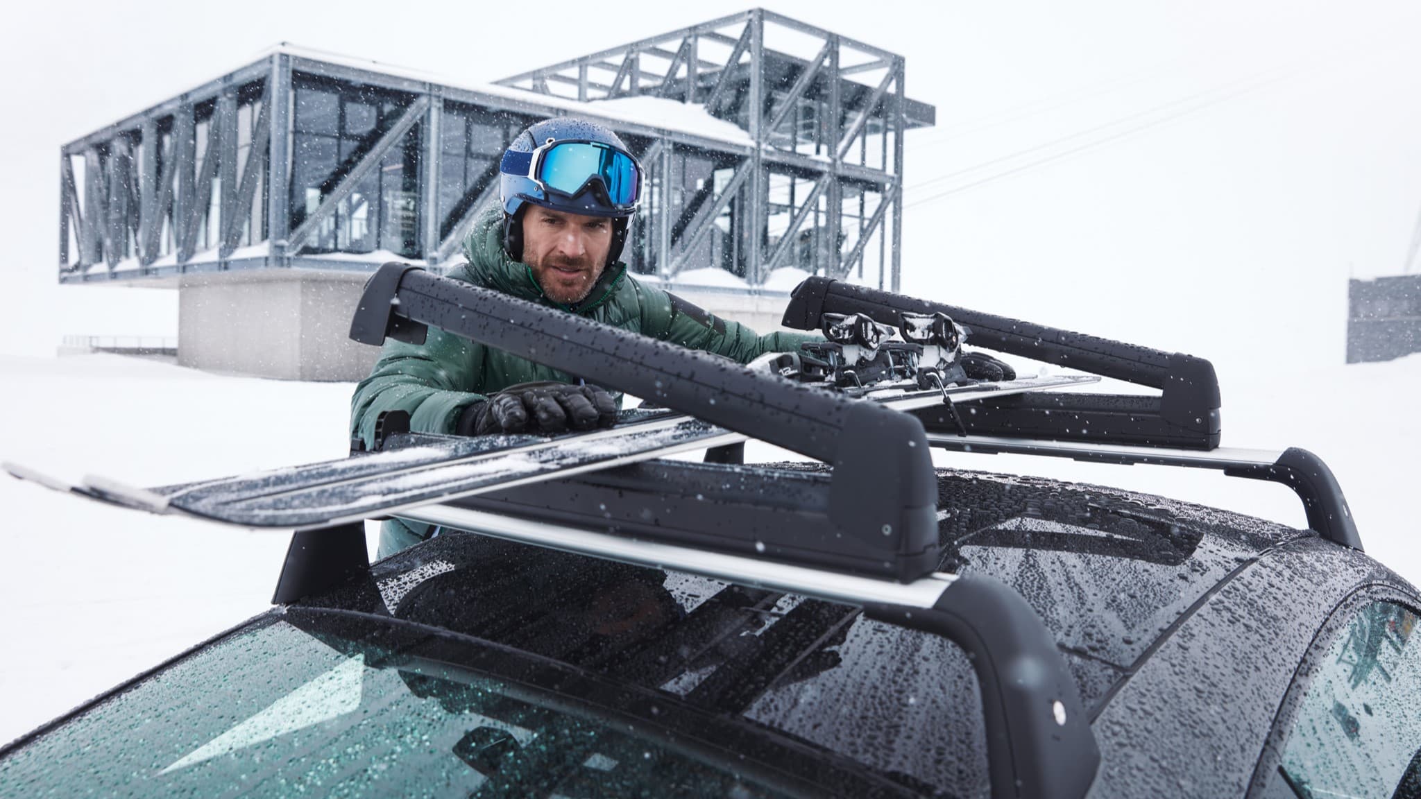 Homme en veste verte et casque bleu fixant des skis sur le porte-bagages de toit d'une voiture dans un paysage enneigé avec un bâtiment moderne en arrière-plan.