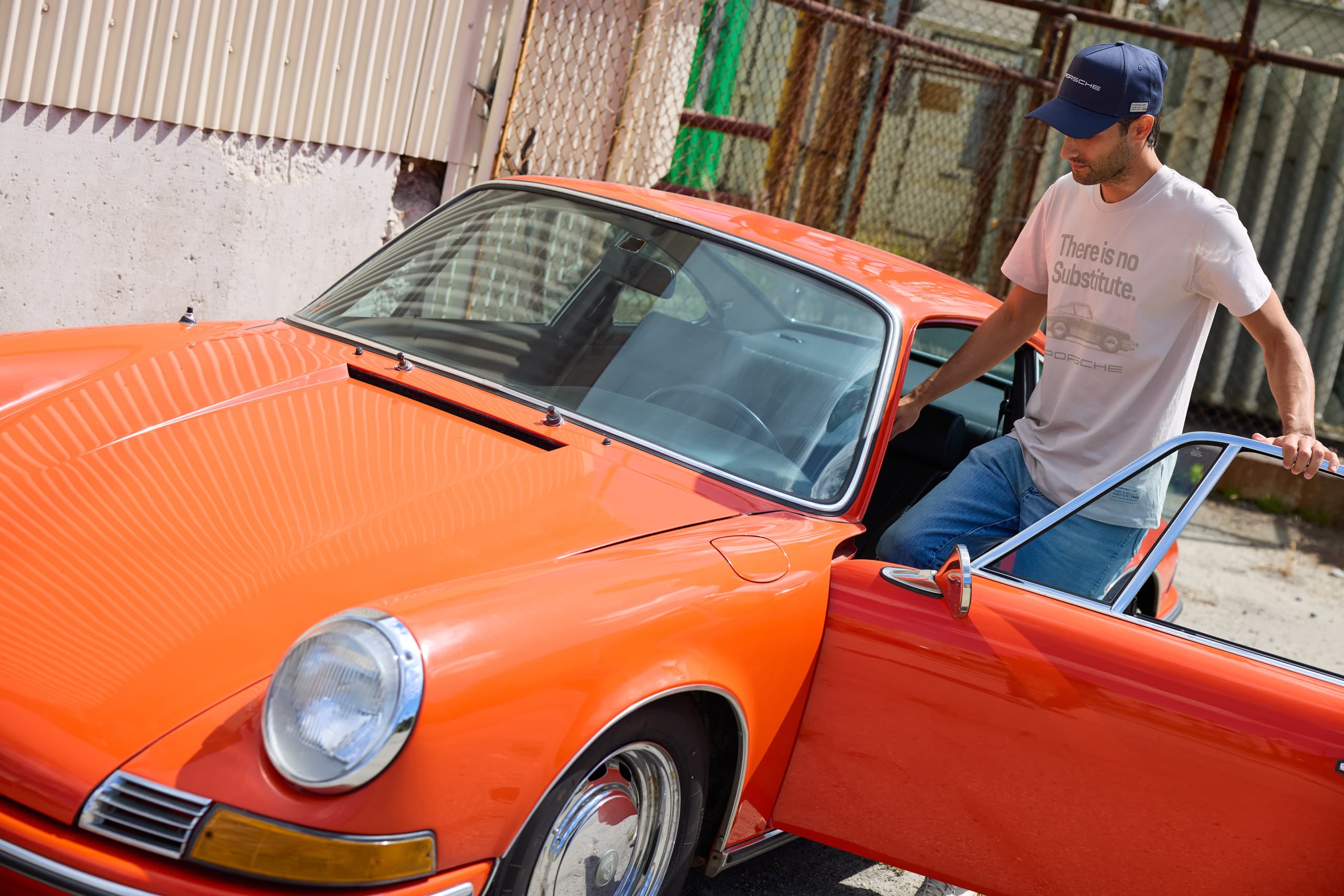 Man with navy Porsche cap and white t-shirt gets into an orange classic Porsche 911.