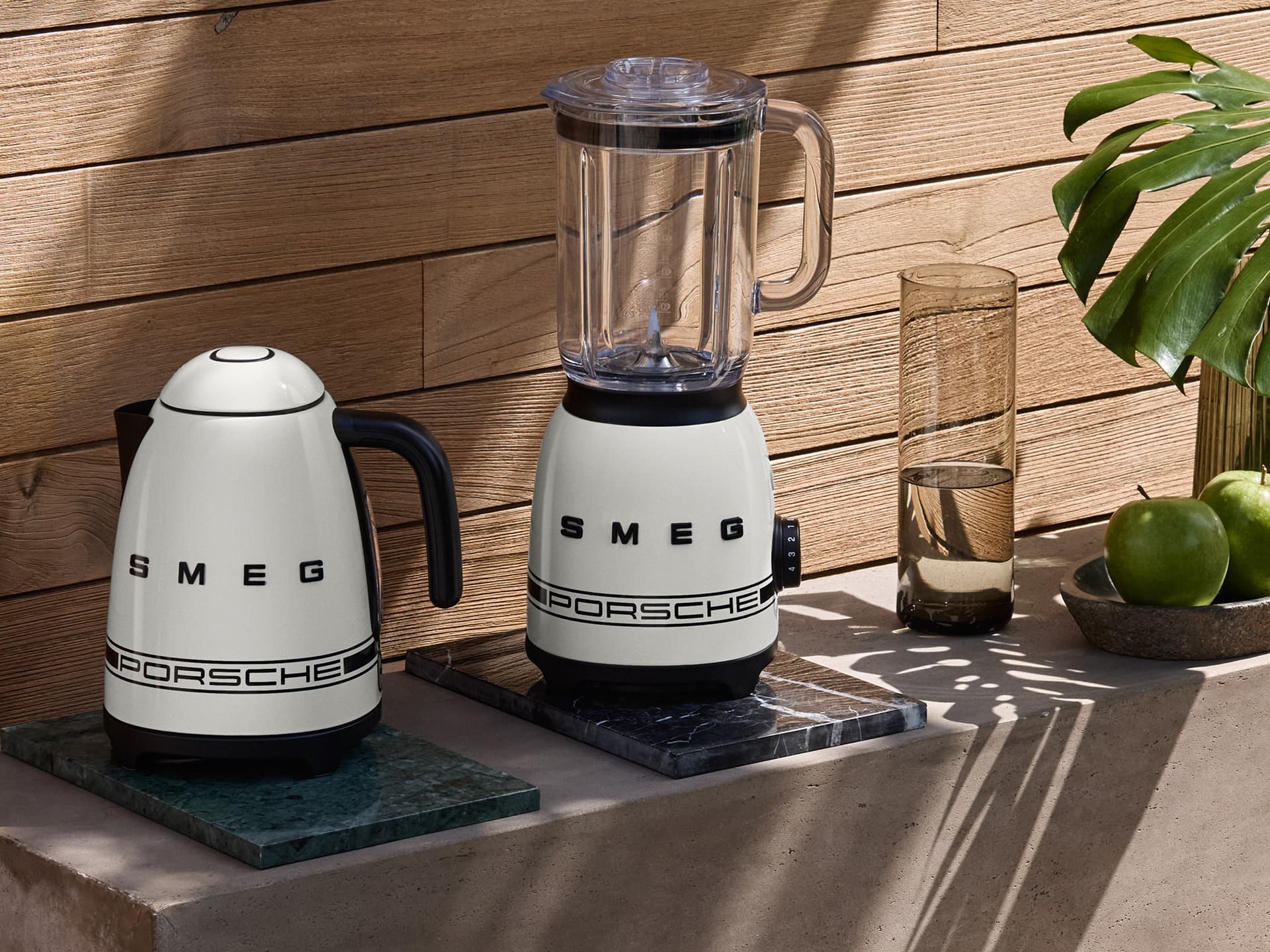 White Smeg kettle and blender with Porsche branding on a wooden counter, next to a glass of water and green apples.