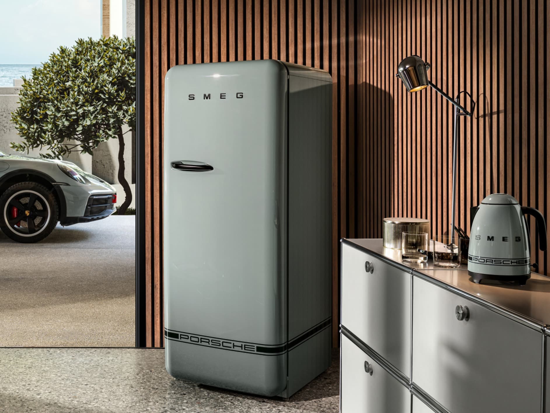 Retro-Kühlschrank und Wasserkocher in Grau mit Porsche-Branding, vor einer Wand aus Holzlatten und moderner Dekoration.