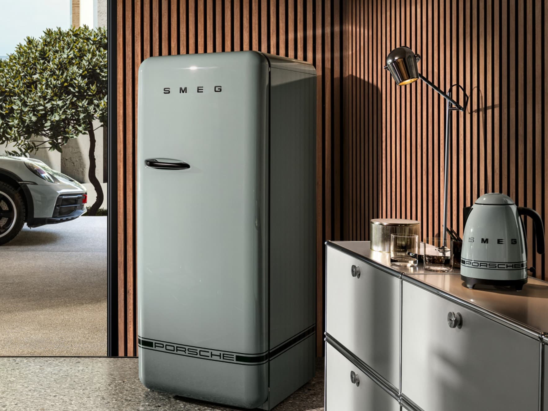 Retro-Kühlschrank und Wasserkocher in Grau mit Porsche-Branding, vor einer Wand aus Holzlatten und moderner Dekoration.