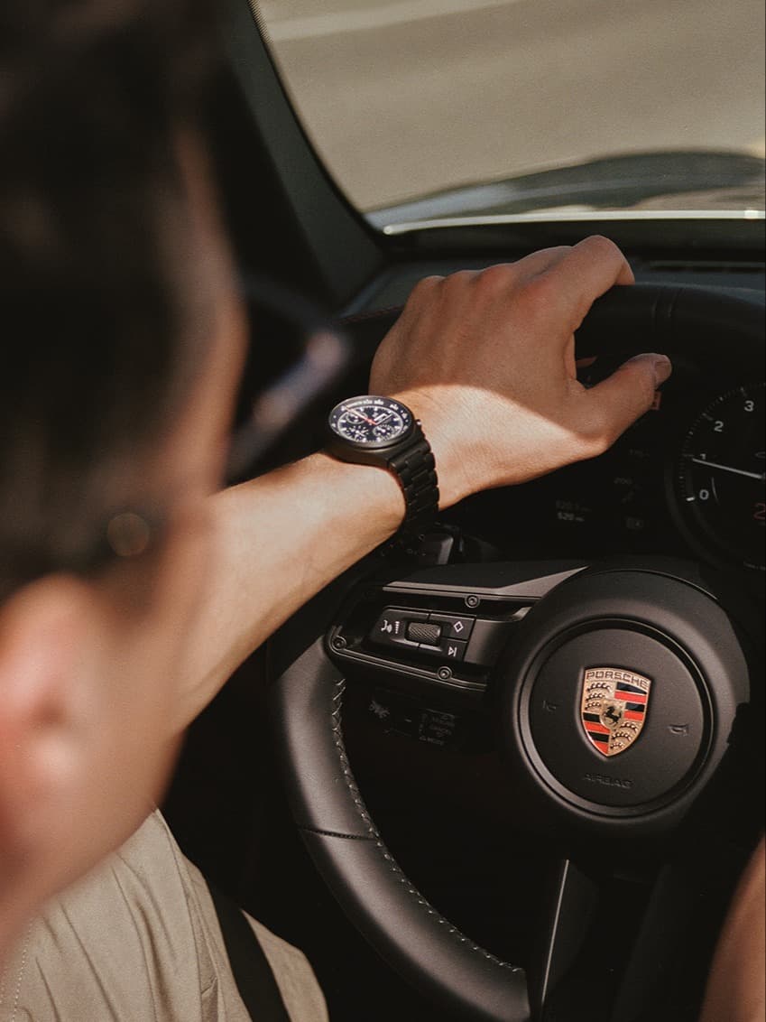 Mano sul volante Porsche, indossa un orologio nero, vista dall'interno dell'auto.