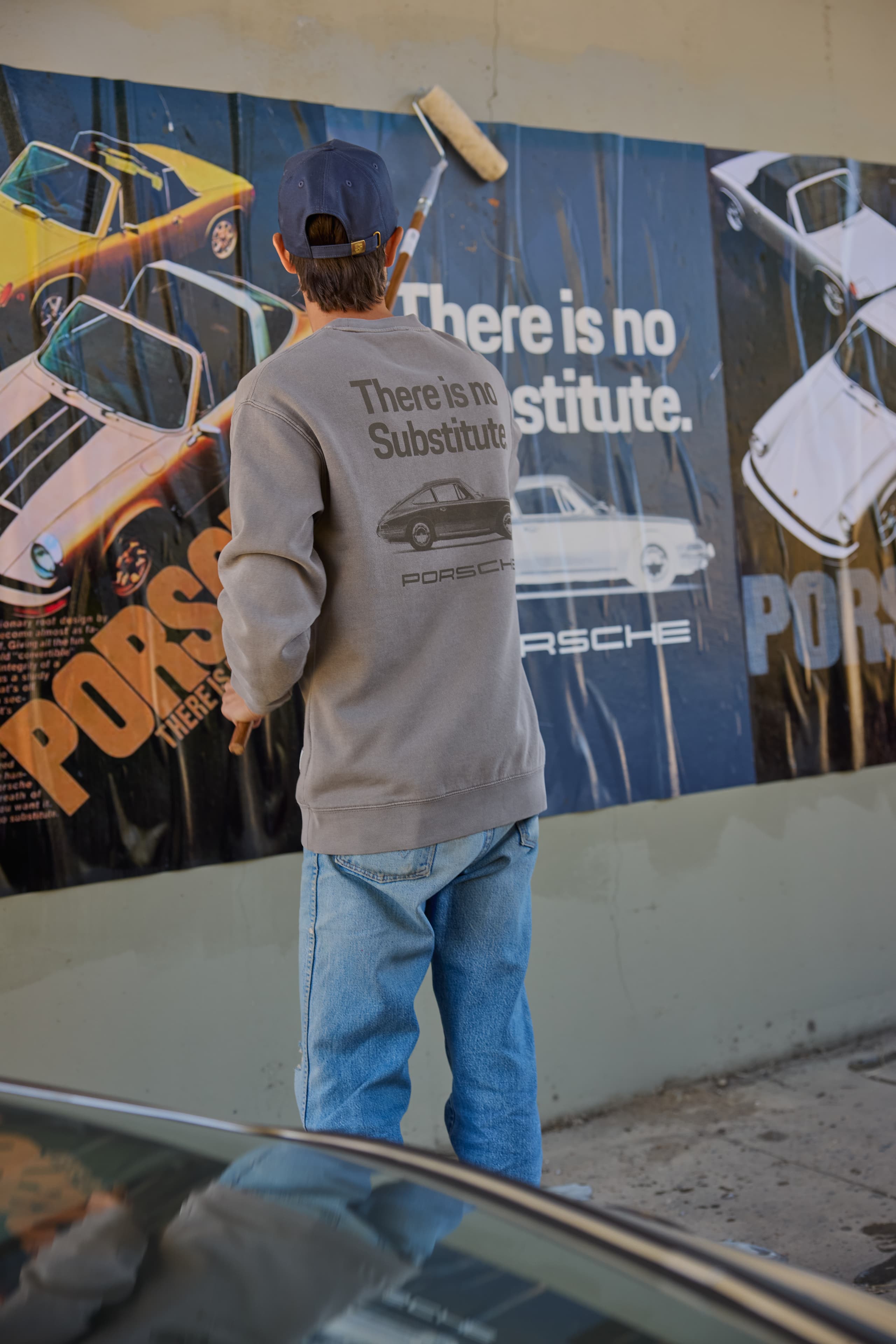 Person in grey Porsche sweatshirt and blue cap pasting a Porsche poster on a wall with a paint roller