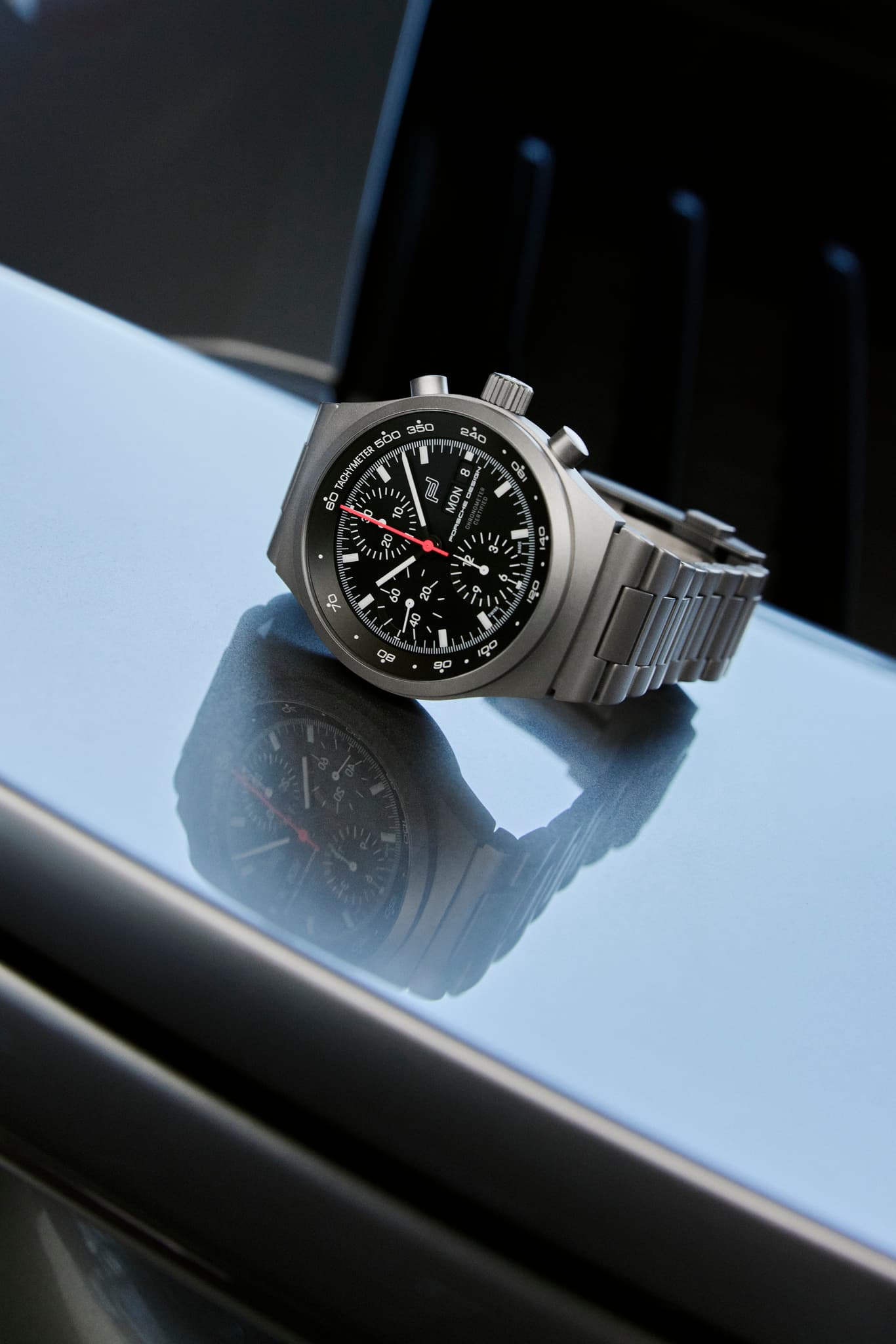 Silberne Chronographen-Armbanduhr mit schwarzem Zifferblatt und rotem Sekundenzeiger, auf glänzender Fläche.