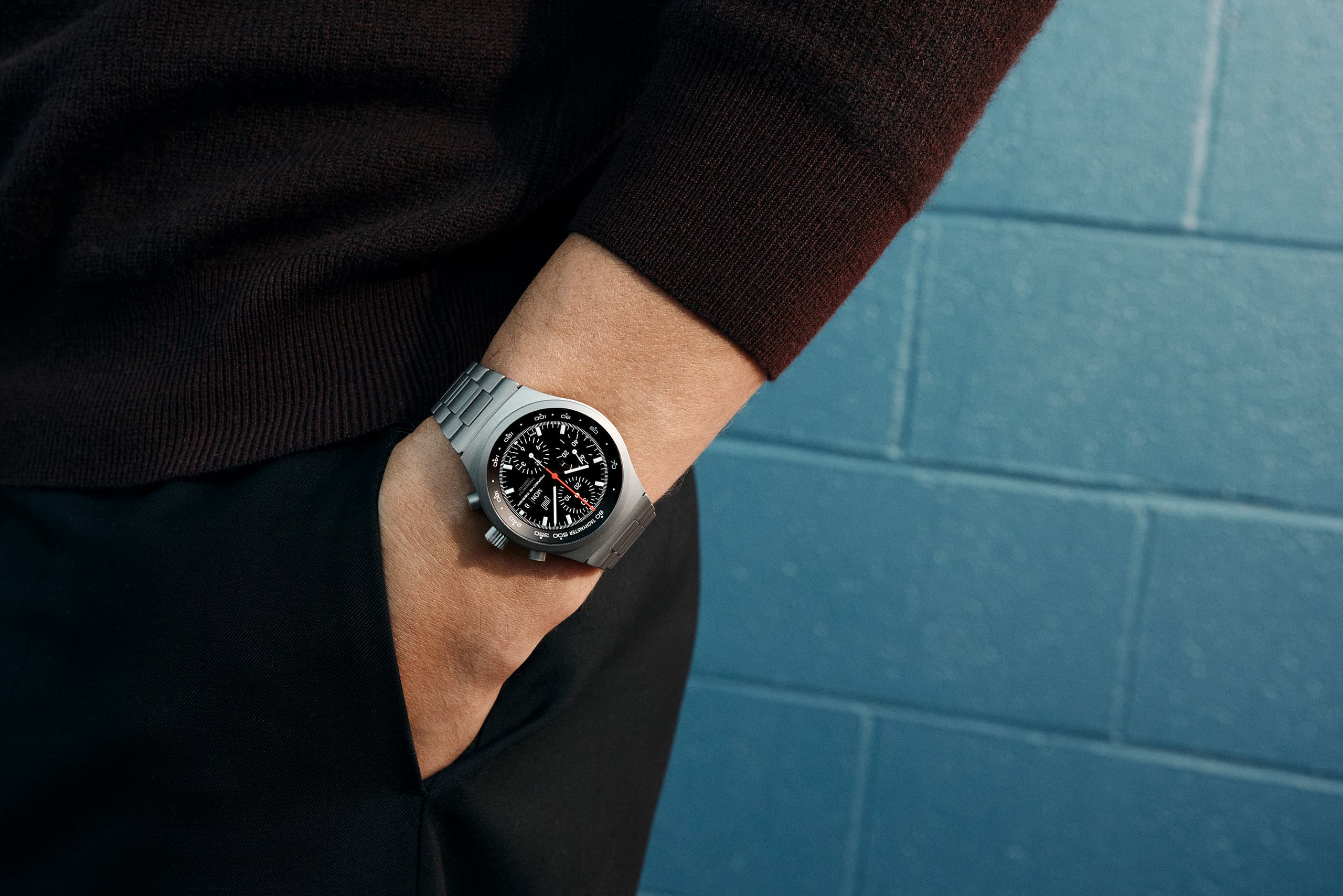 Silver chronograph watch with black dial on a wrist, worn with a dark sweater against blue tiles