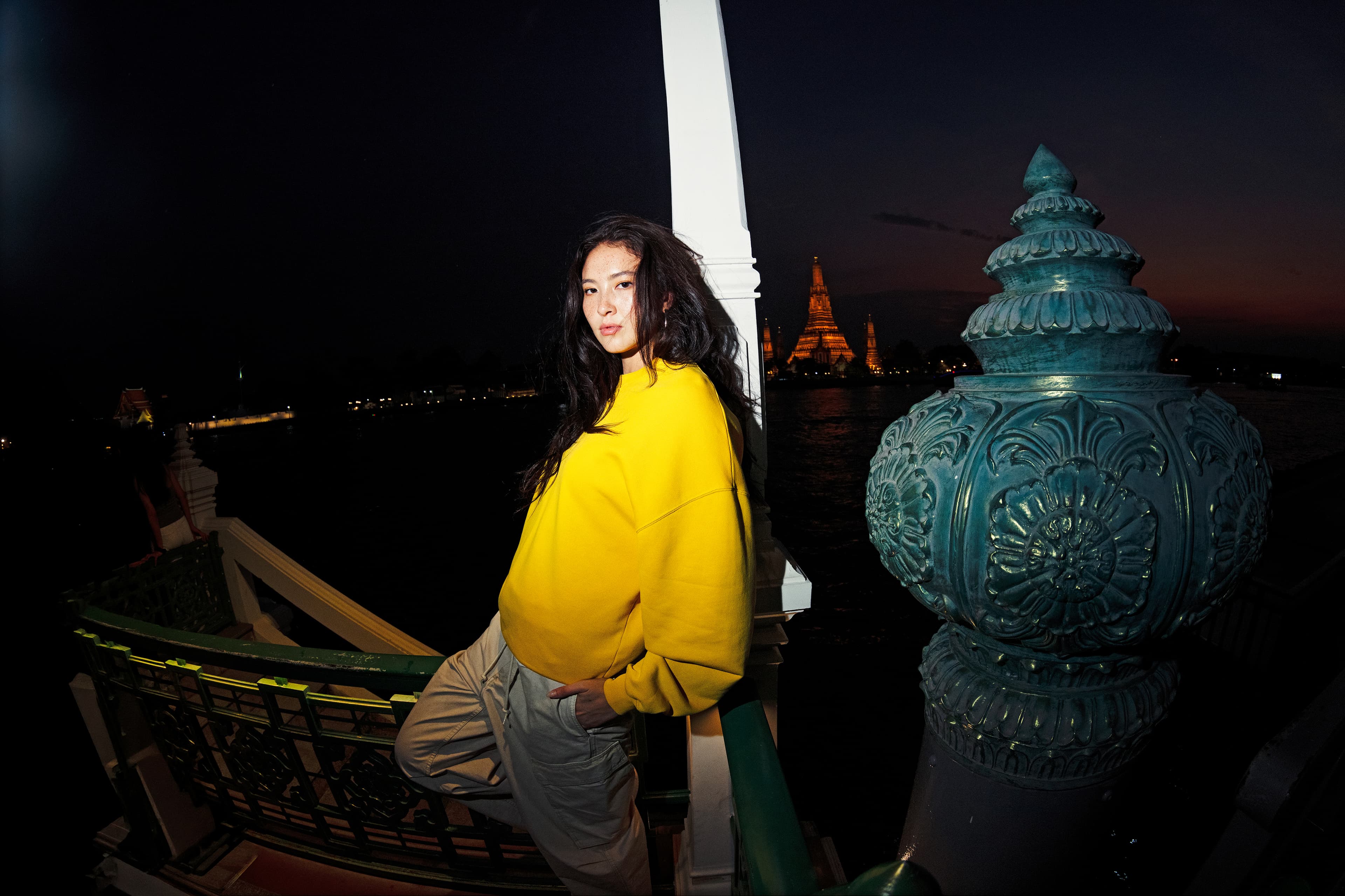Woman wearing a yellow Porsche AHEAD sweatshirt and beige pants on riverside steps at night, temple lights in the background.