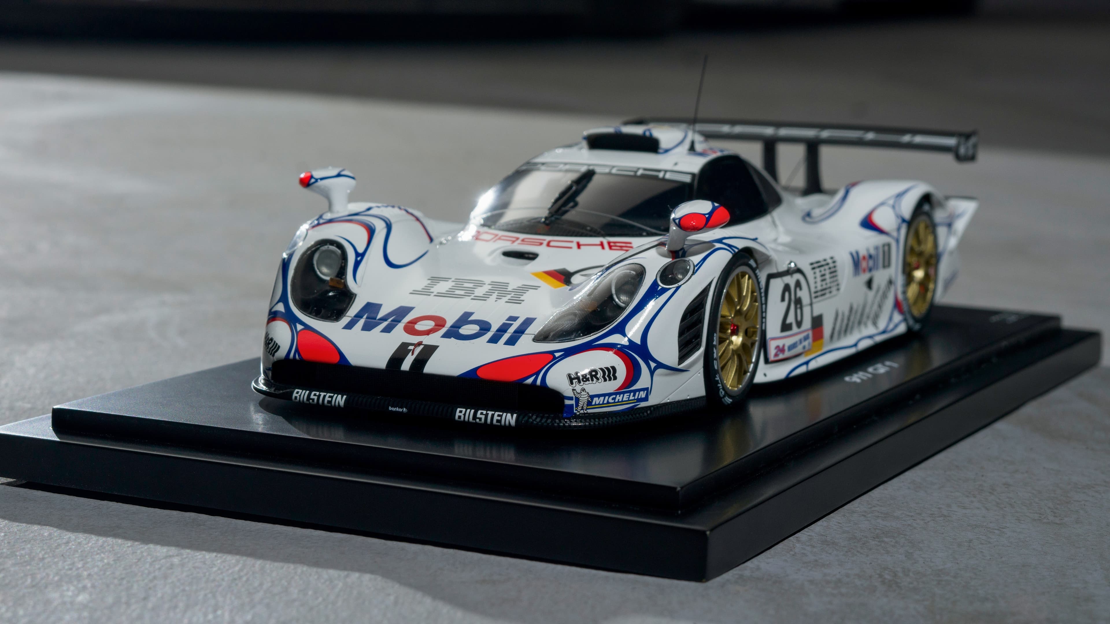 Model van een witte Porsche raceauto met kleurrijke decals en gouden wielen op een zwart displaystandaard.
