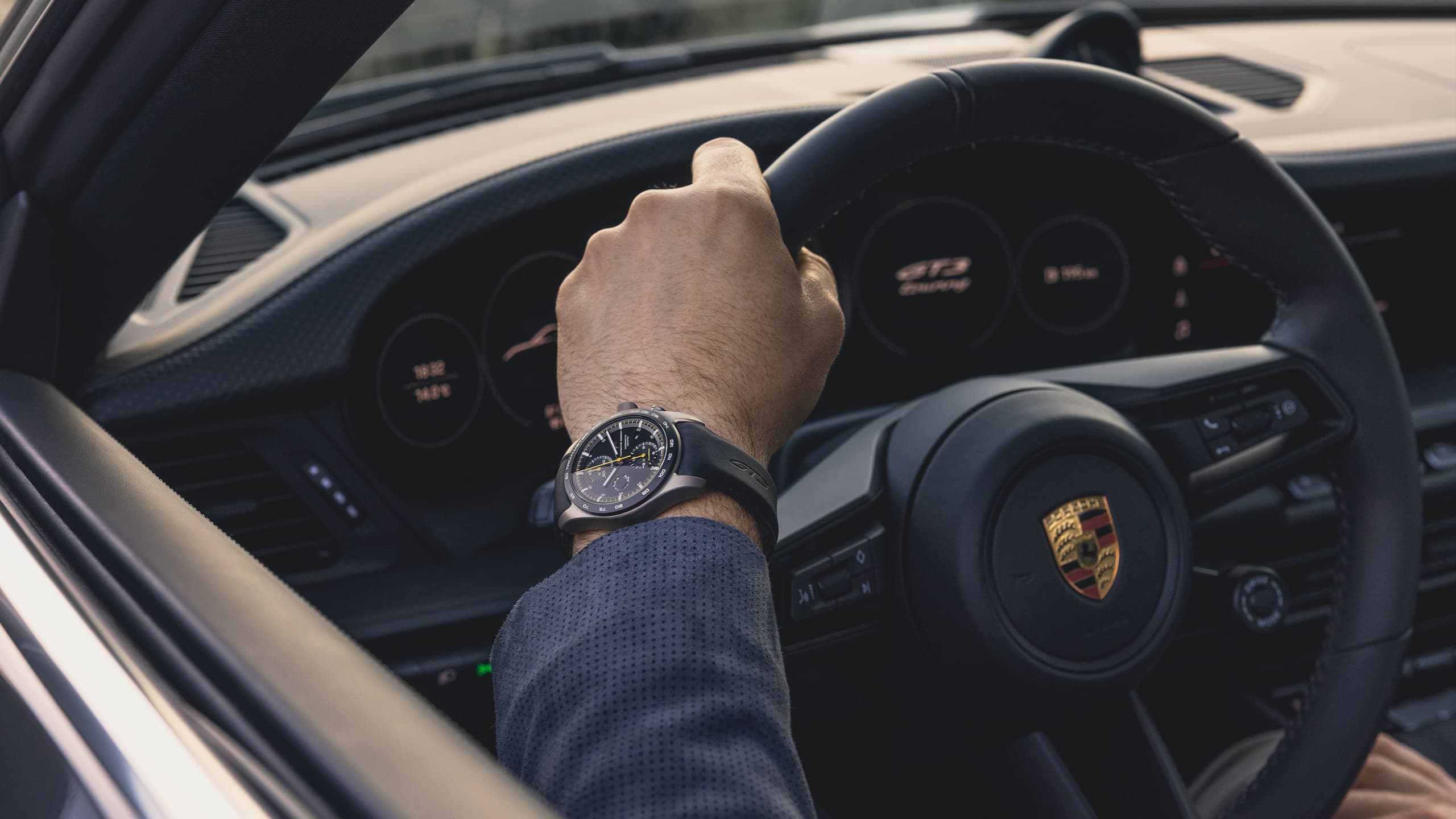 Dłoń na kierownicy Porsche, na nadgarstku czarny smartwatch z okrągłą tarczą, we wnętrzu kokpitu.