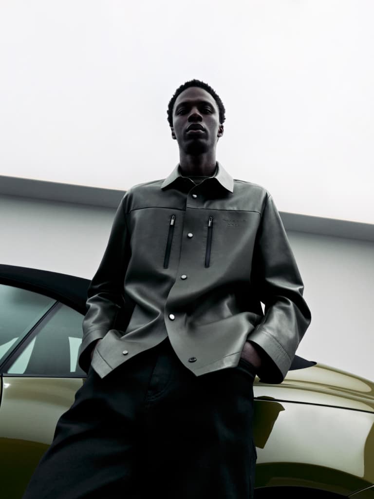 Homme en veste grise se tient avec assurance à côté d'une voiture verte, sous un ciel nuageux.