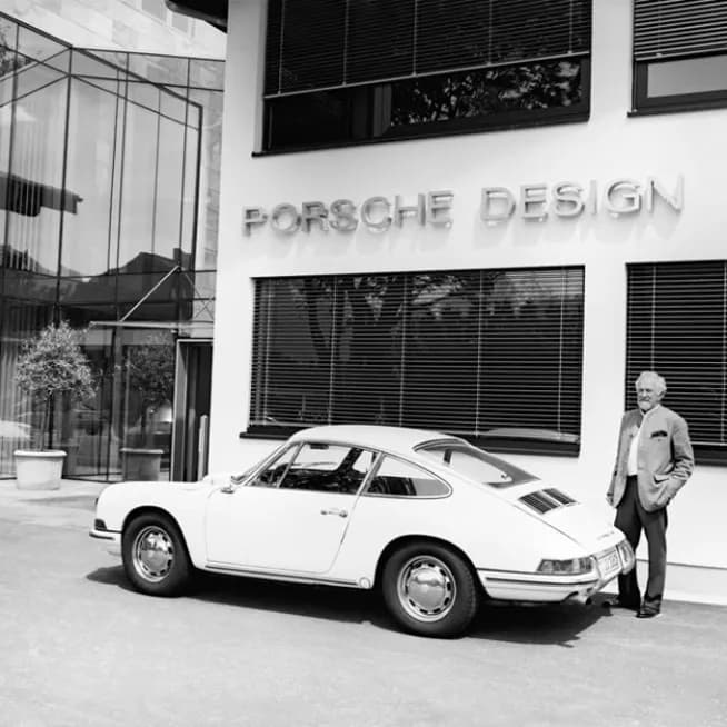 Czarno-białe zdjęcie: biały Porsche 911 przed budynkiem z napisem „Porsche Design”, obok stoi mężczyzna.