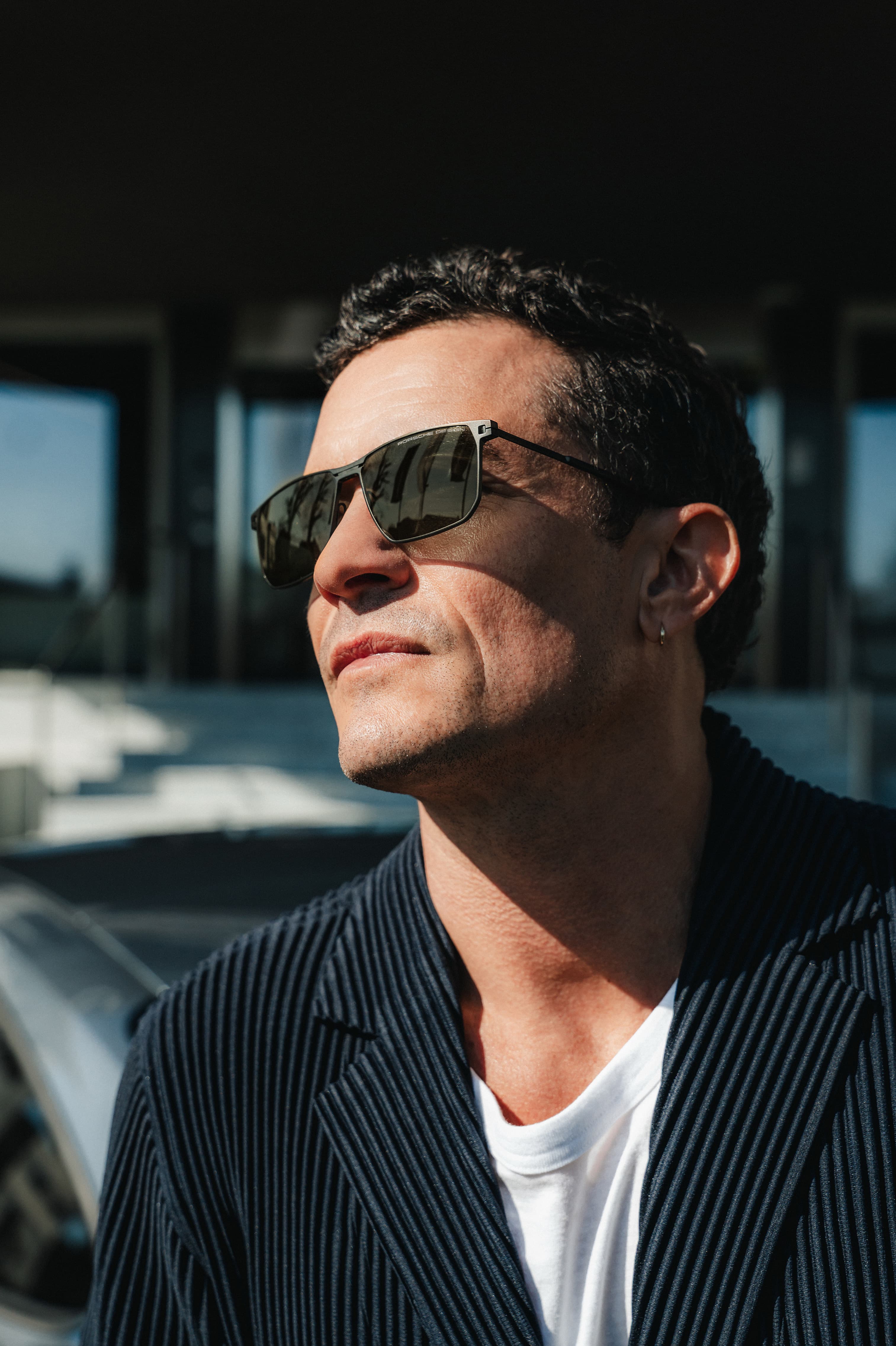 Orlando Bloom trägt schwarze Sonnenbrille von Porsche Design und eine dunkelblaue Strickjacke im Sonnenlicht, urbaner Hintergrund.