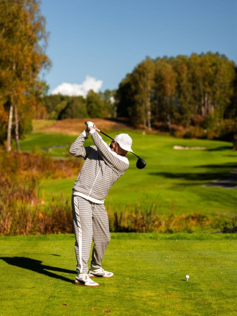 Golfer wearing a white cap and a checkered outfit teeing off on a green fairway, with trees and a blue sky in the background.