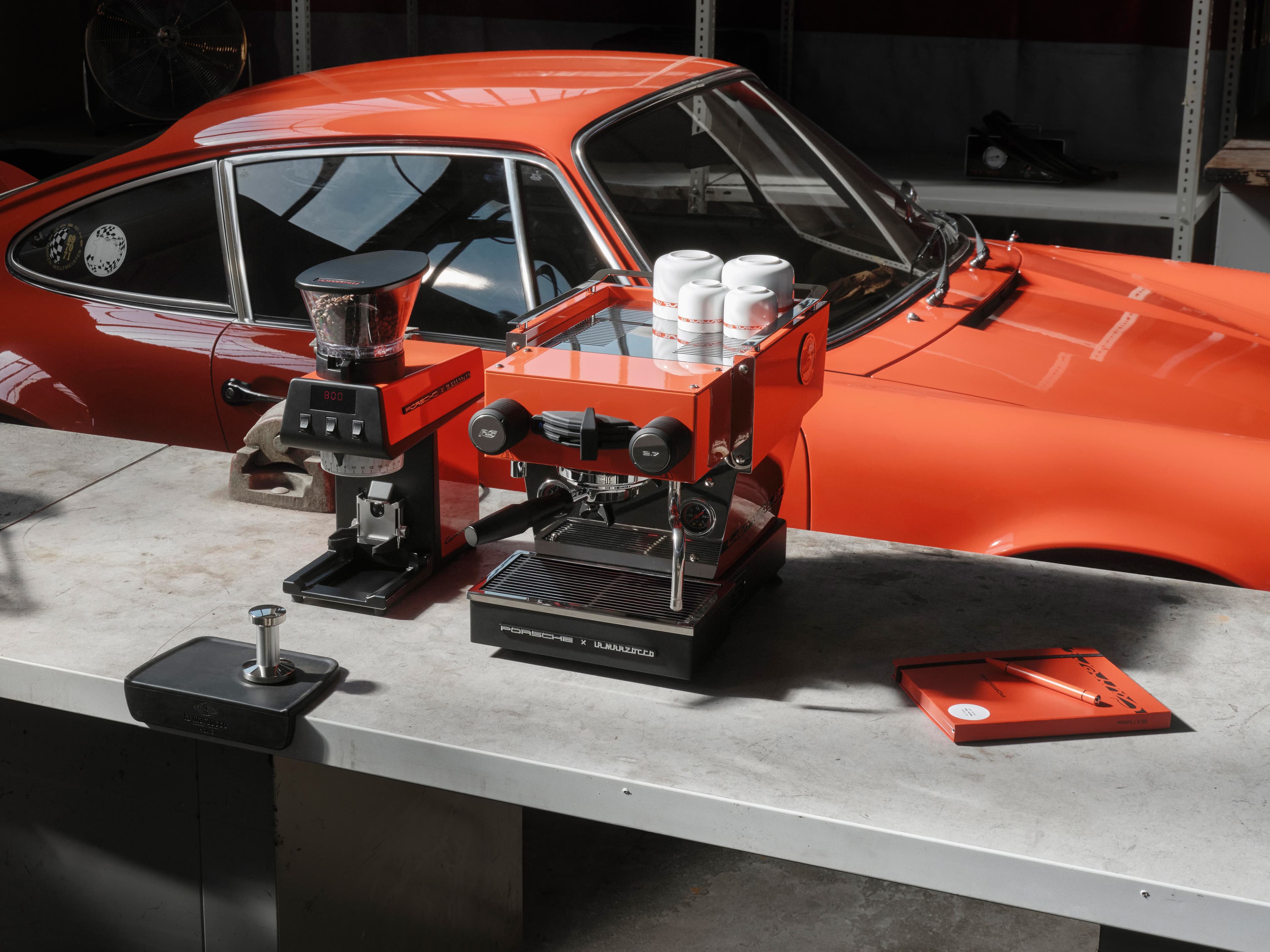Machine à expresso et moulin à café La Marzocco x Porsche avec porte-filtre, posés sur un établi, à côté d’une Porsche 911 Carrera RS 2.7 dans un atelier.