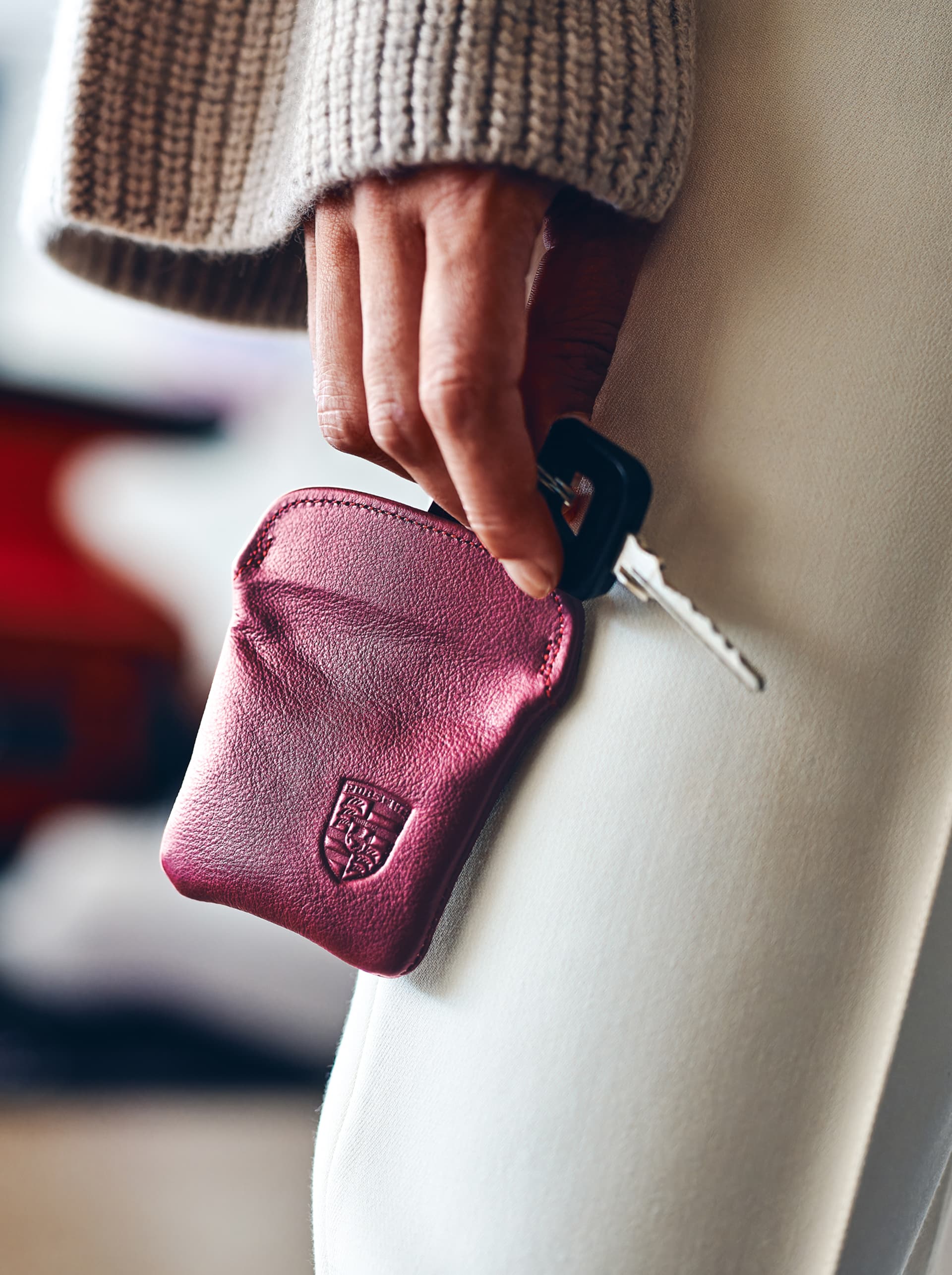 Hand holding a car key with a burgundy leather key pouch, embossed with a logo, against a light background.