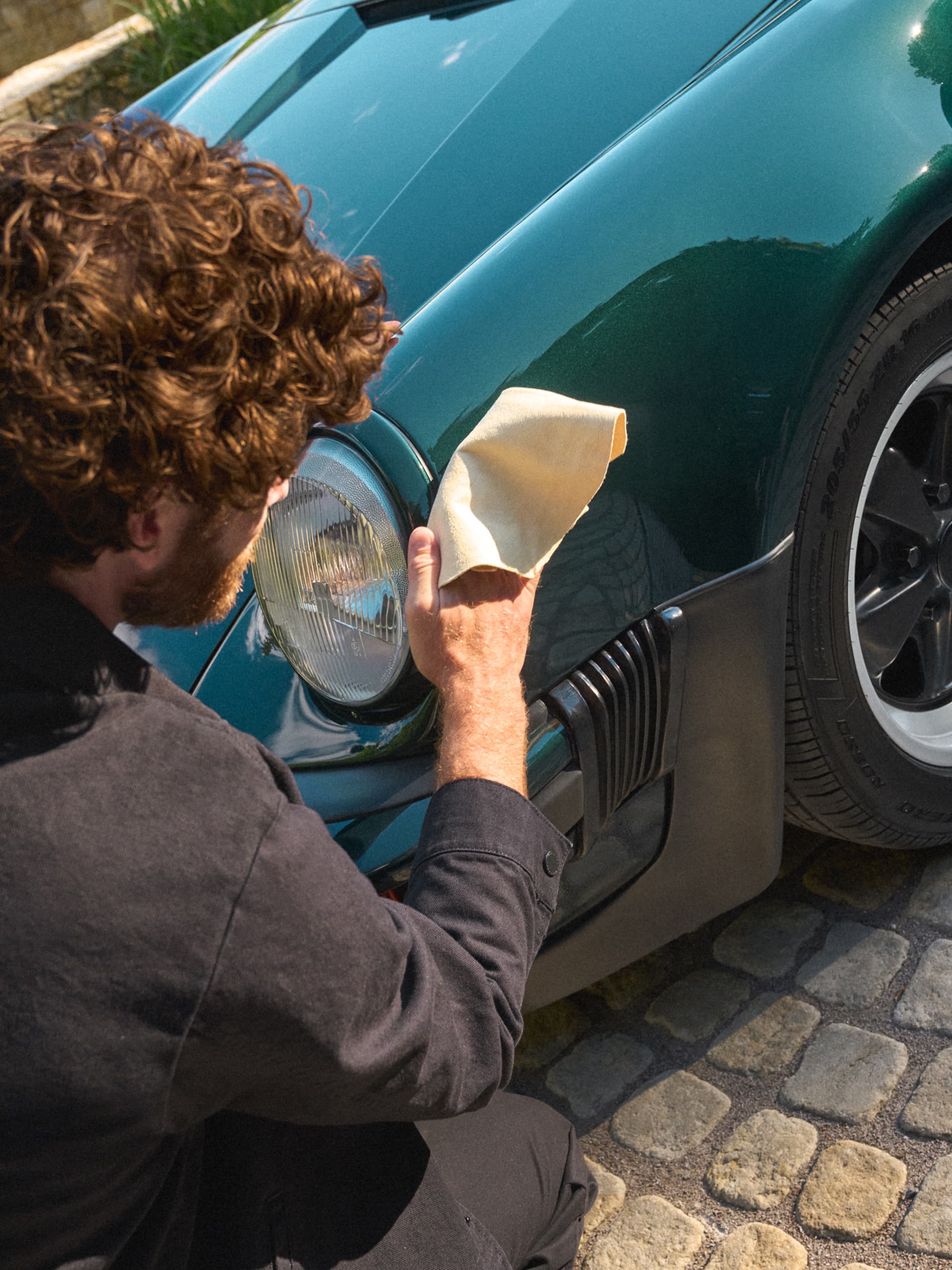 Person polishing the front fender of a dark green Porsche with a beige cloth, close-up by the headlight and wheel.