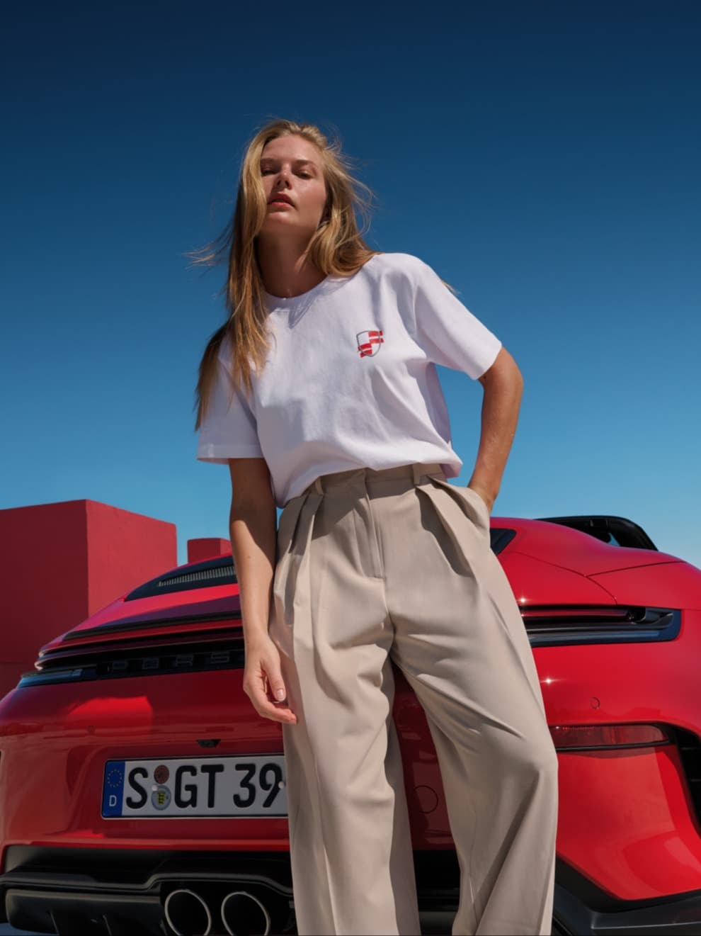Femme en T-shirt blanc et pantalon beige, appuyée devant un cabriolet Porsche rouge sous un ciel bleu.