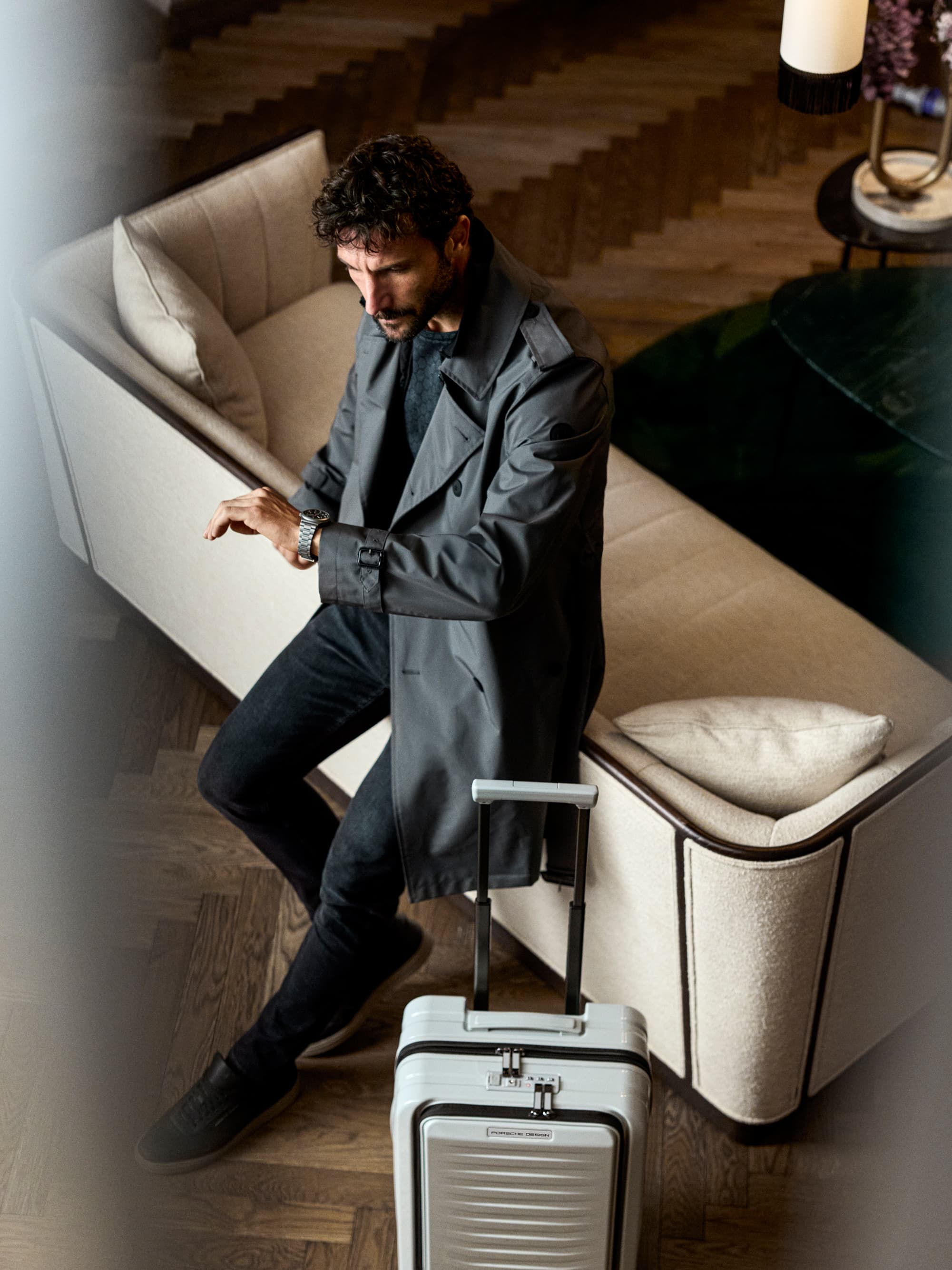Man in gray coat sitting on sofa, checking watch, with silver suitcase on wooden floor.