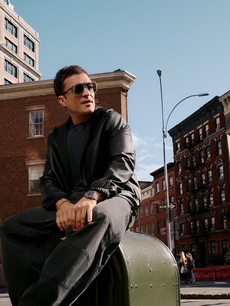 Homme en lunettes de soleil et veste noire assis sur une boîte aux lettres verte dans un cadre urbain, avec des bâtiments en briques et un ciel dégagé en arrière-plan.