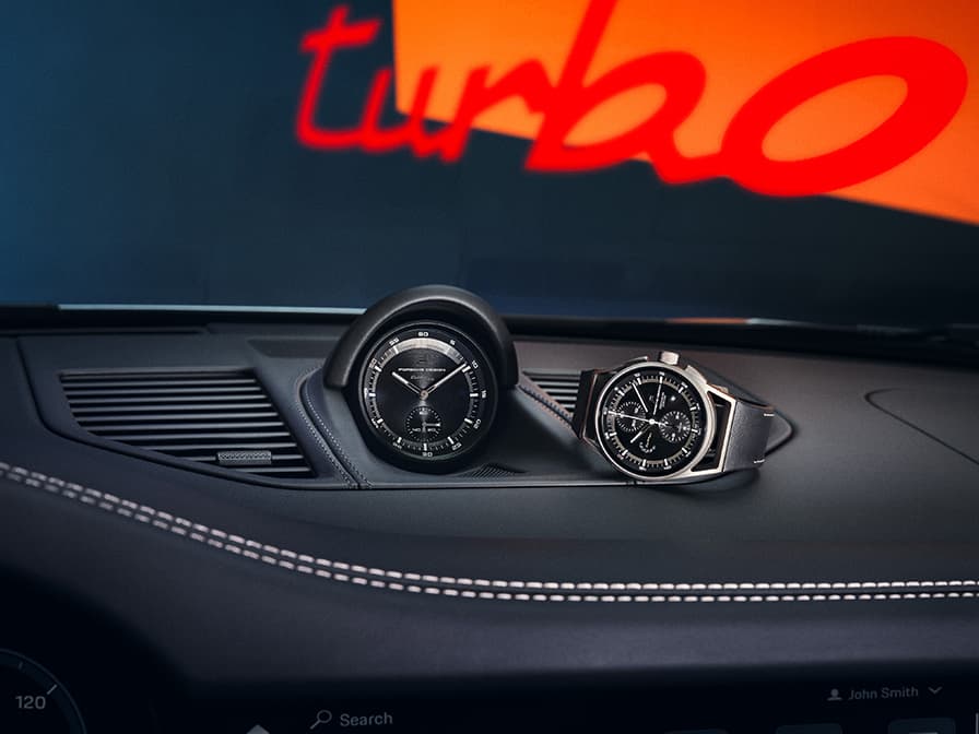 Czarny chronograf Porsche Design obok zegara Sport Chrono na czarnej desce rozdzielczej, w tle napis „turbo”.