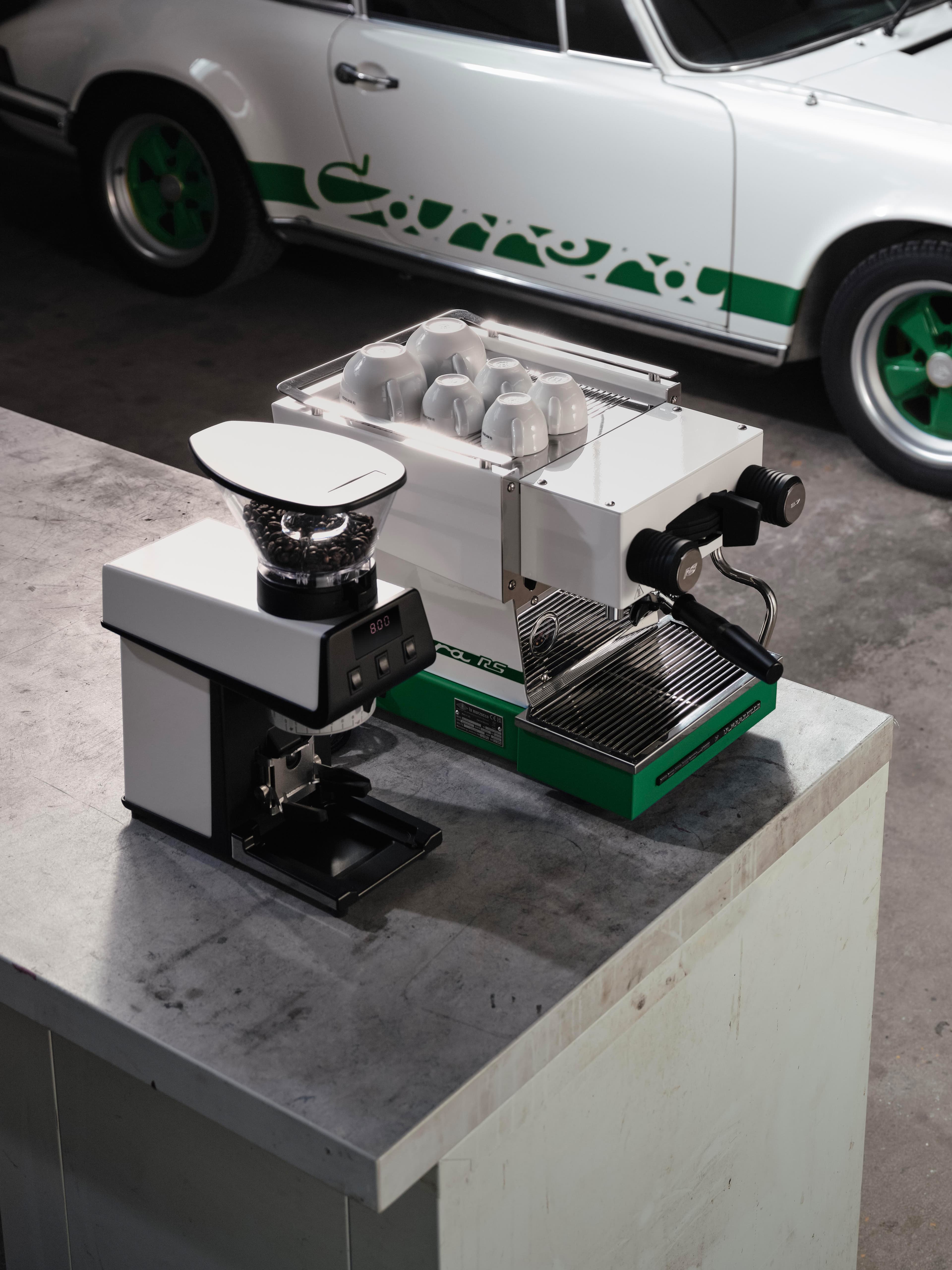 Ekspres do espresso i młynek do kawy na betonowym bloku, w tle białe Porsche z zielonym logo Carrera.