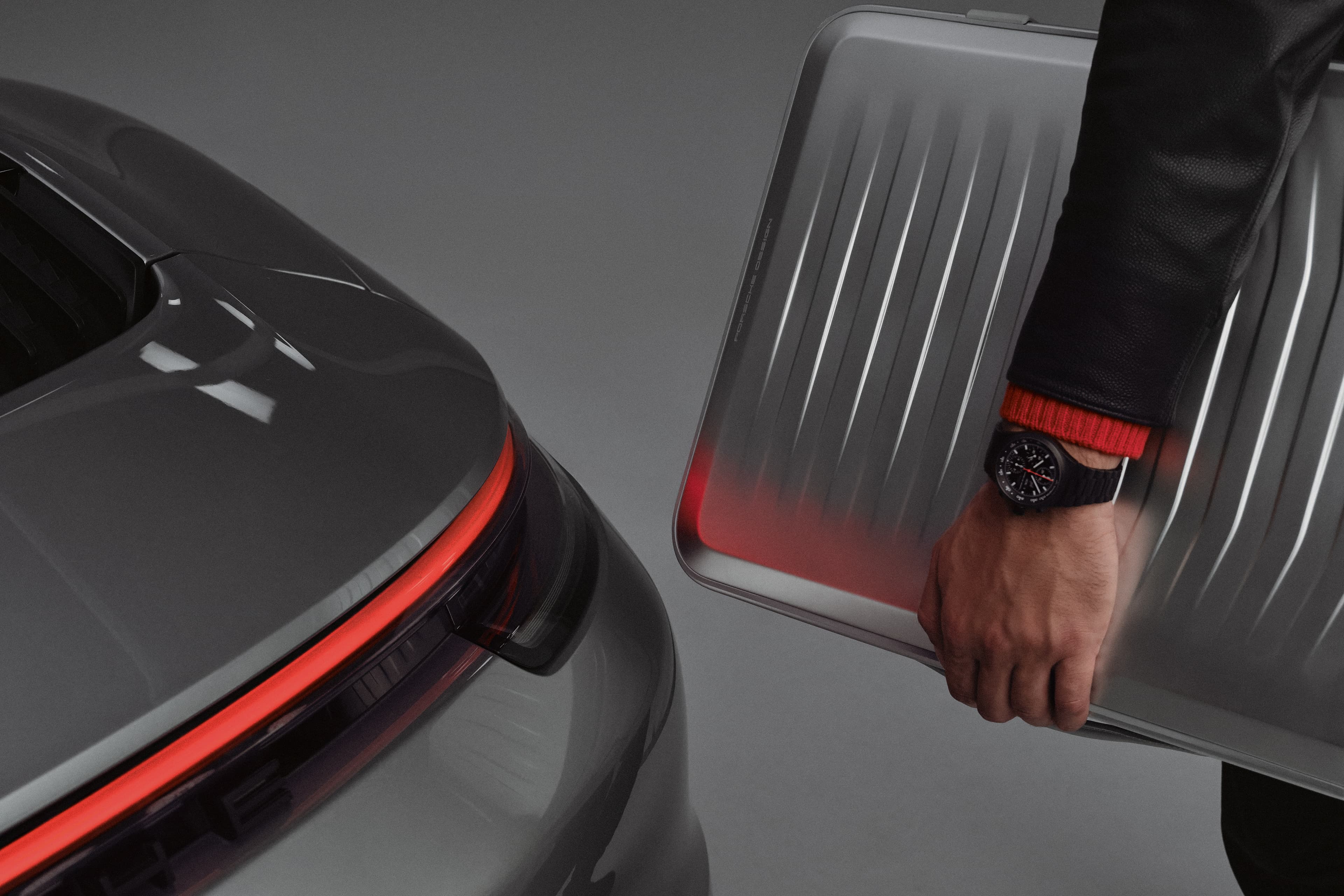 Hand holding silver Porsche Design suitcase beside a grey Porsche rear with red taillight strip.