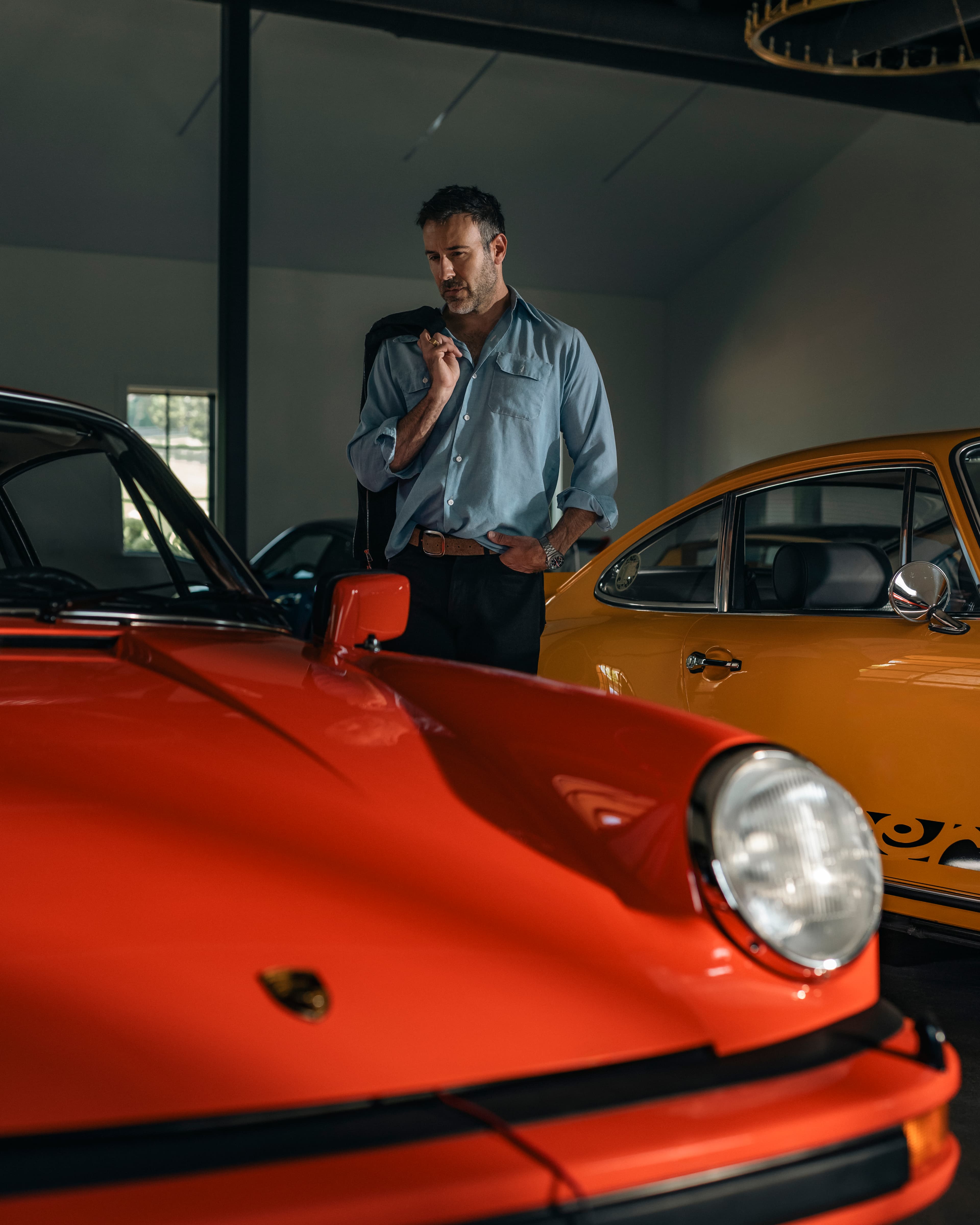 Uomo con giacca nera appoggiato a un’auto d’epoca gialla in un garage, altre auto sportive sullo sfondo.