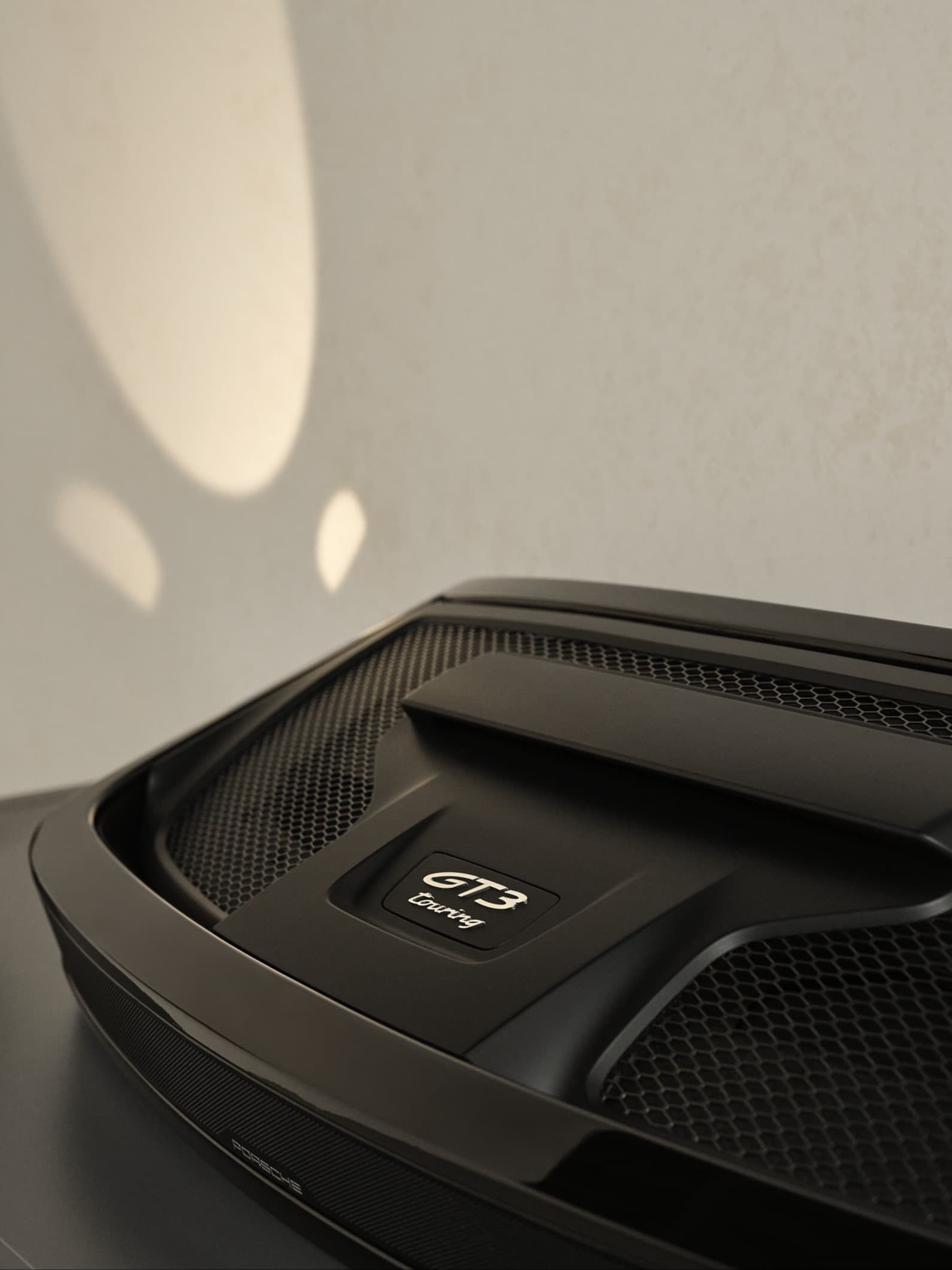 Gros plan du capot moteur noir de la Porsche GT3 Touring avec détails en maille, sur fond de mur clair avec ombres douces.