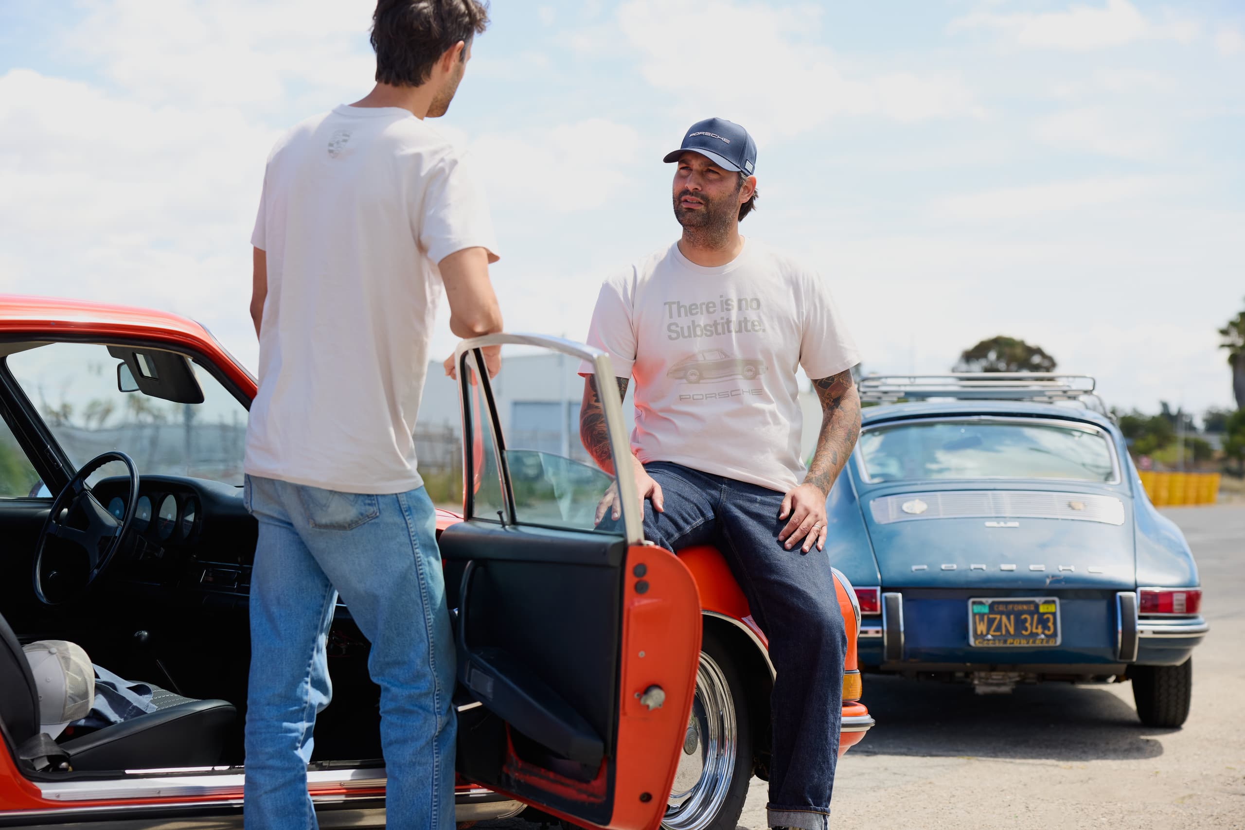 Two men in white Porsche T-shirts by an orange classic car, with a blue Porsche parked behind under a sunny sky.