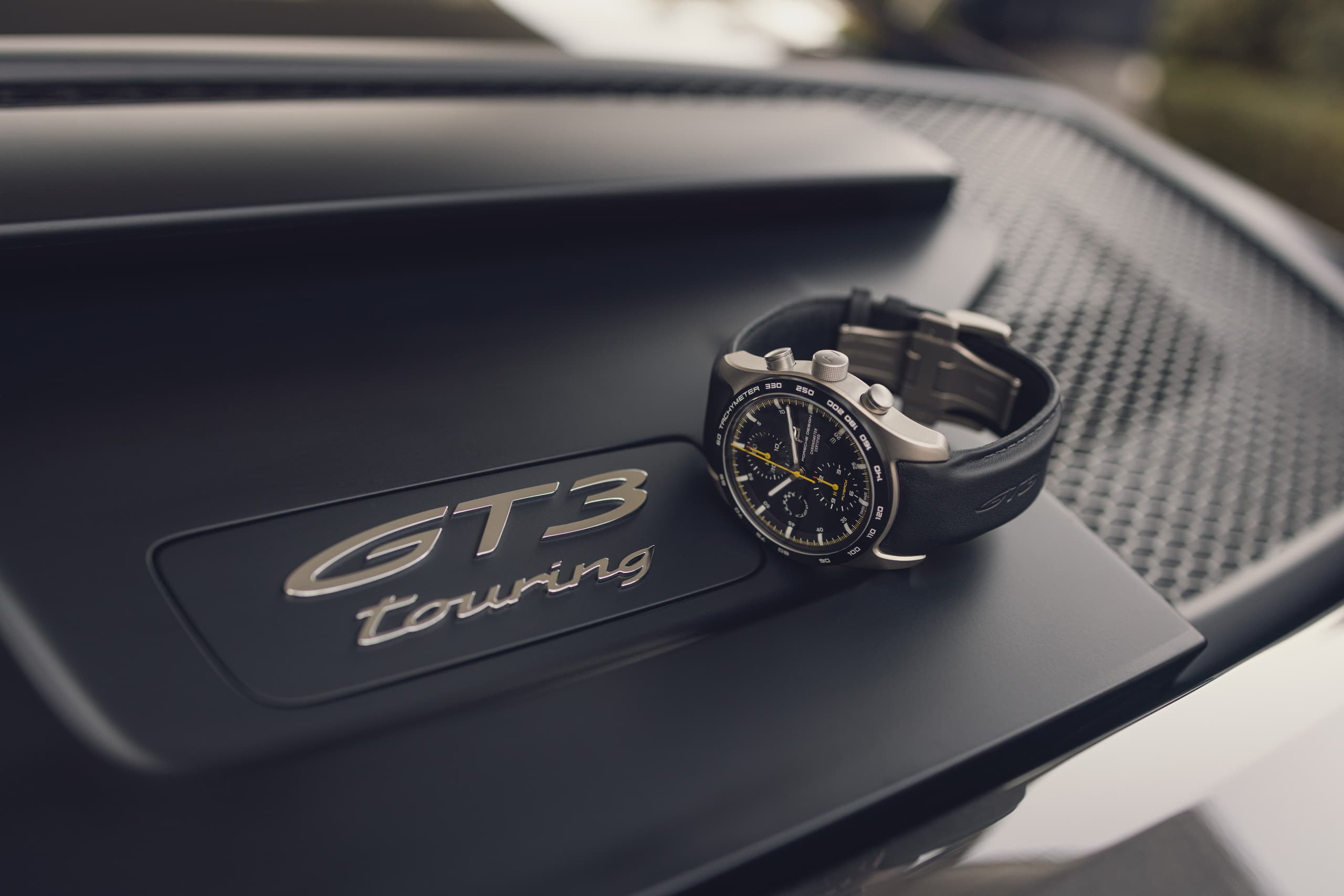 Zegarek chronograf z czarnym skórzanym paskiem, spoczywający na pokrywie silnika Porsche 911 GT3 Touring.
