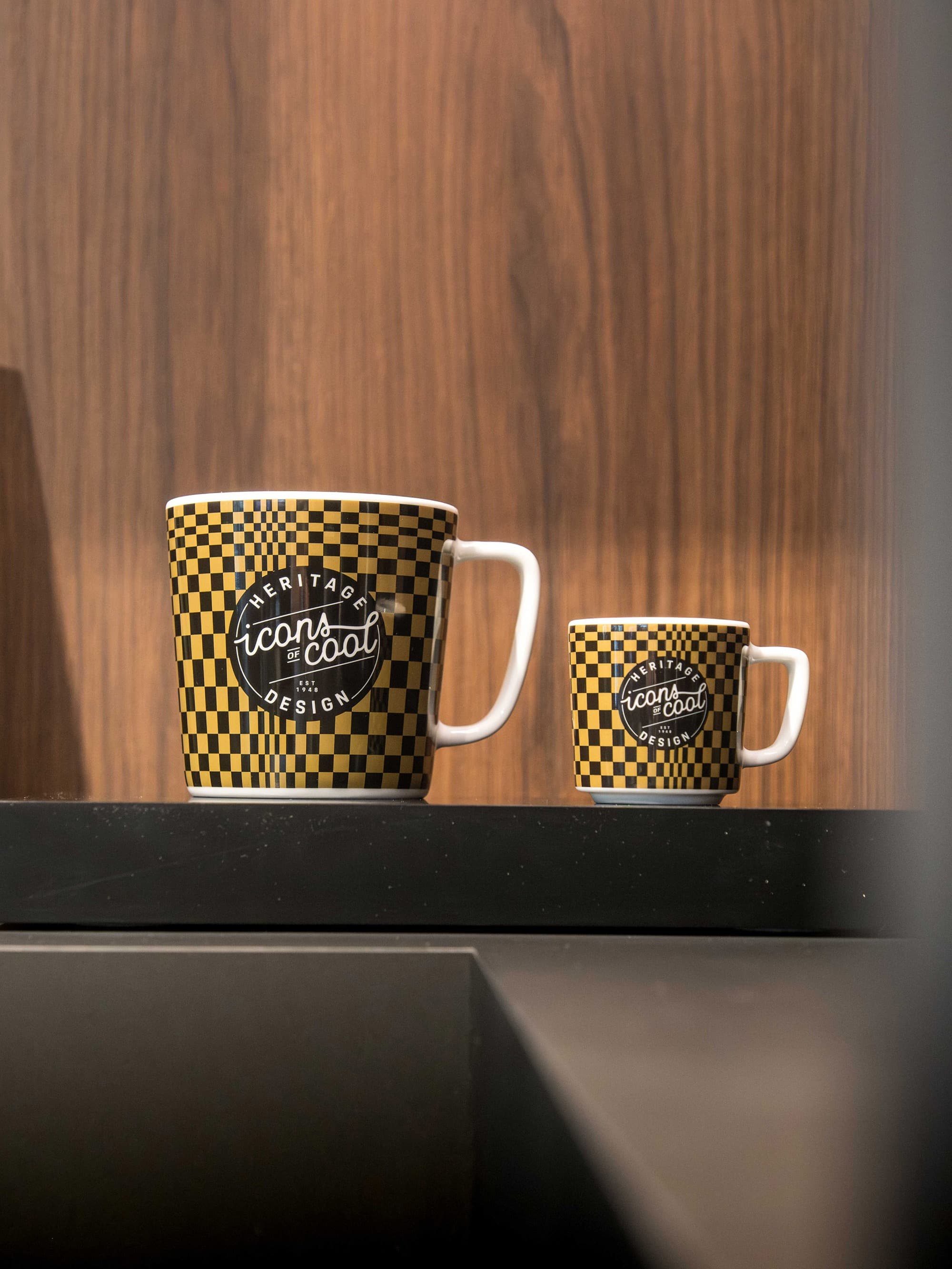 Two ceramic mugs with a black-and-yellow check pattern and “Icons of Cool” logo on a black shelf in front of a wood-paneled wall.