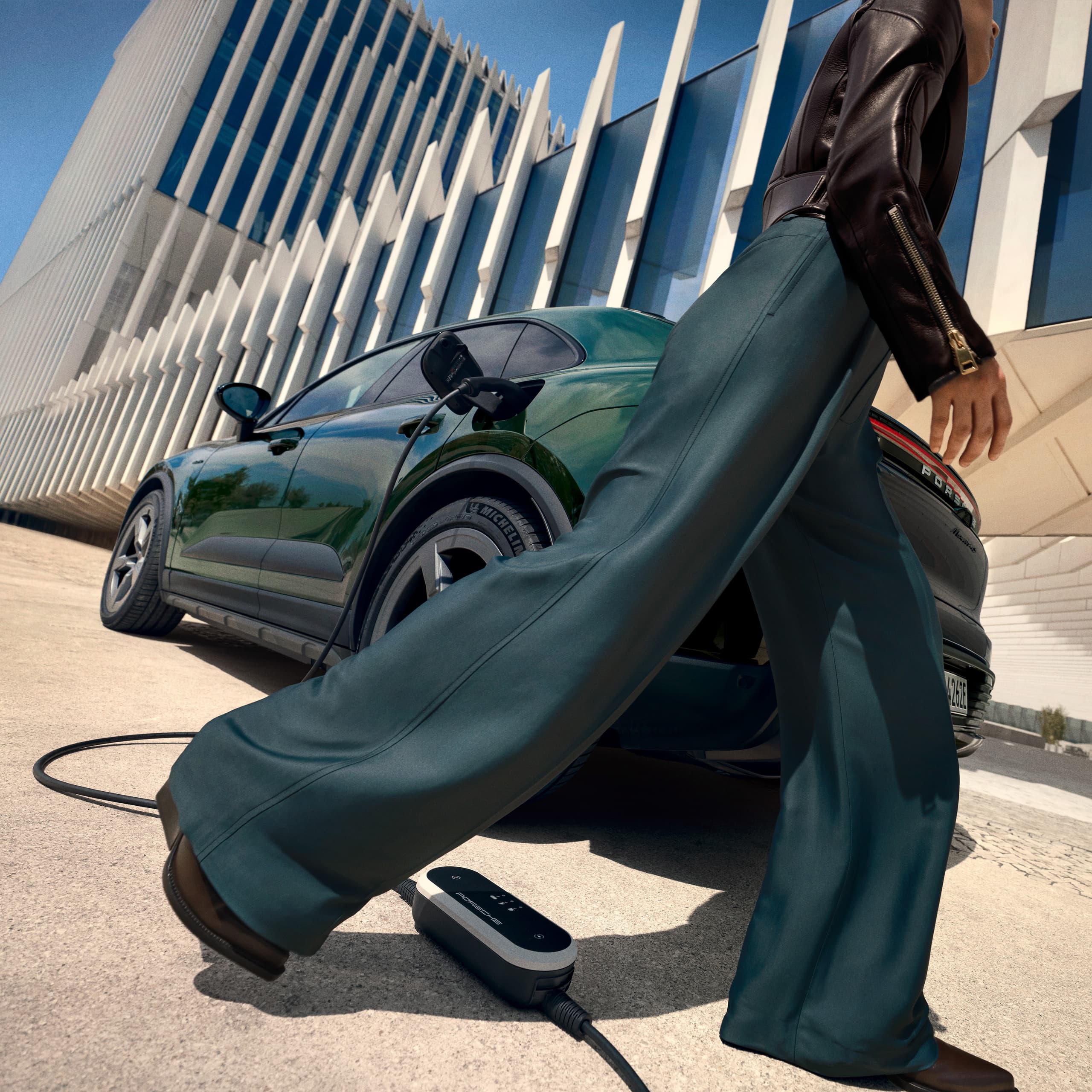 Woman is walking in front of a darkgreen Porsche Macan with a mobile charger on the ground, modern building in the background