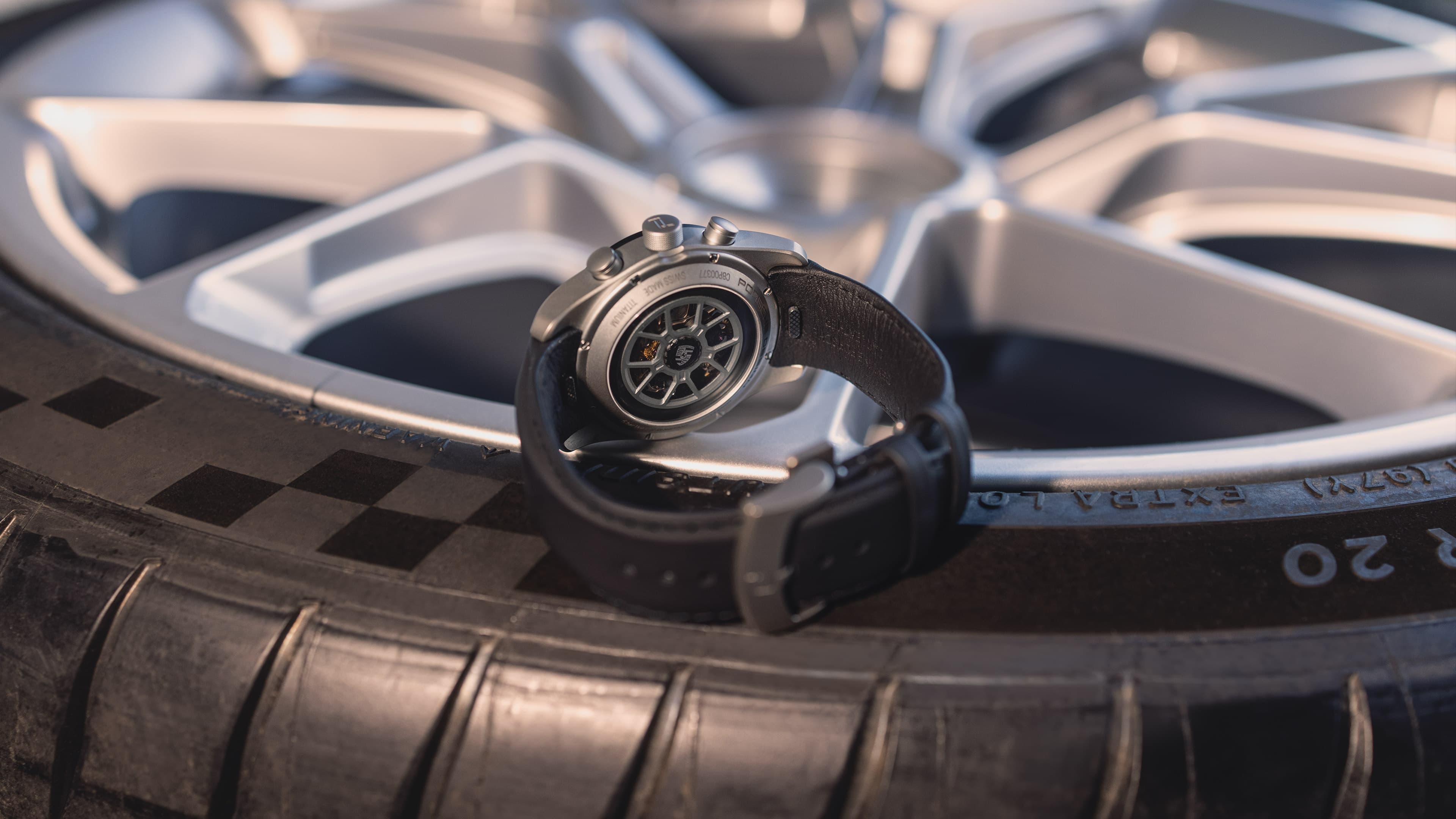 A black wristwatch with a leather strap on a car tire, with a silver rim in the background.