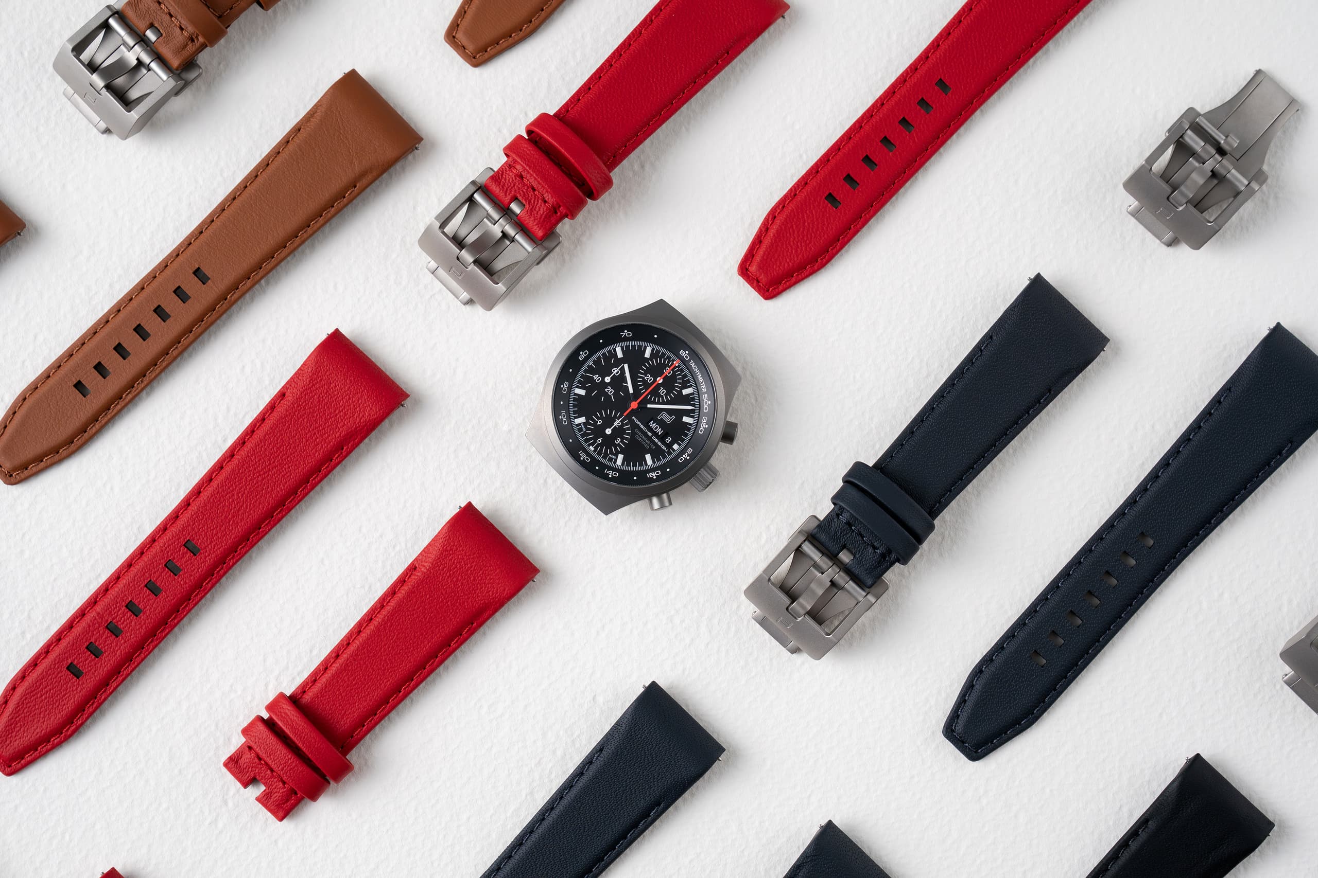 Orologio cronografo con quadrante nero, circondato da cinturini intercambiabili in pelle rossi, marroni e blu navy su sfondo bianco.