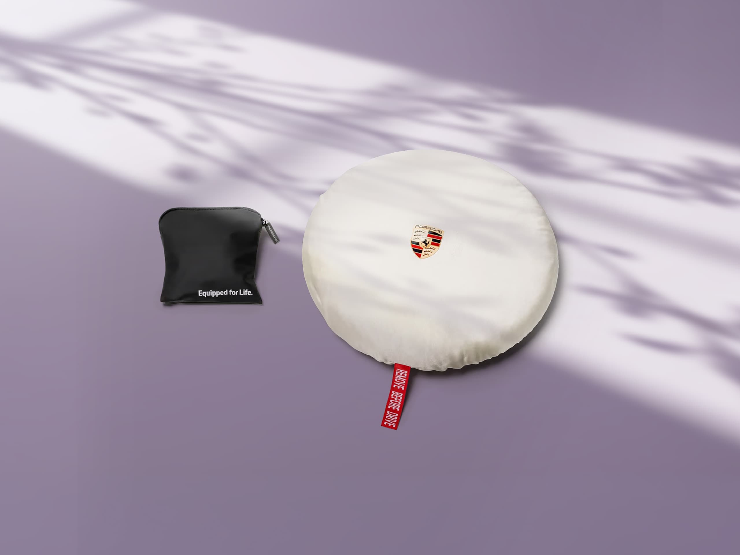 Capa de pneu Porsche branca com logótipo e etiqueta vermelha, ao lado de uma bolsa preta sobre um fundo roxo com padrões de sombra.