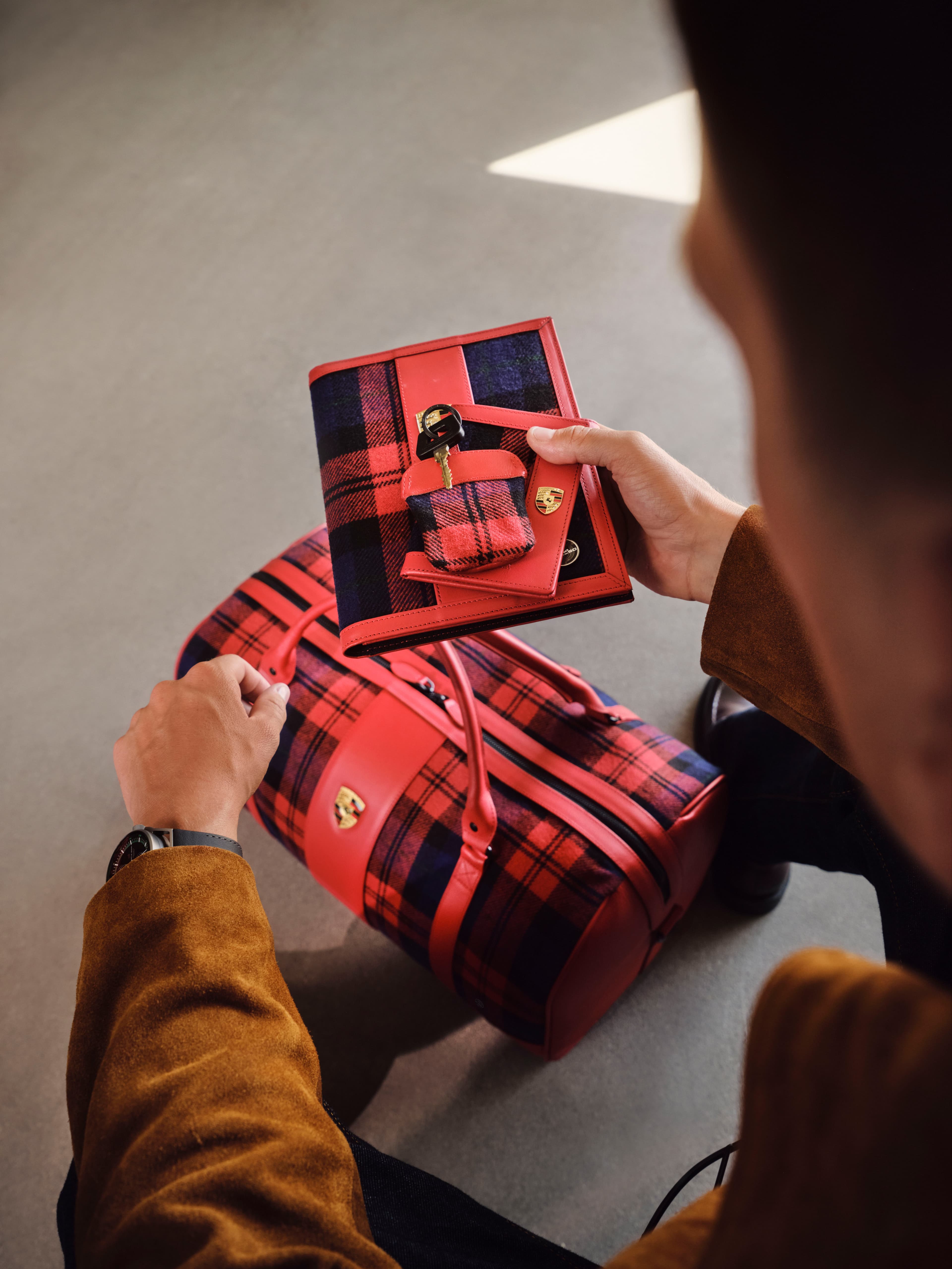 Person holding red and navy tartan Porsche notebook with key pouch, above matching tartan travel bag.