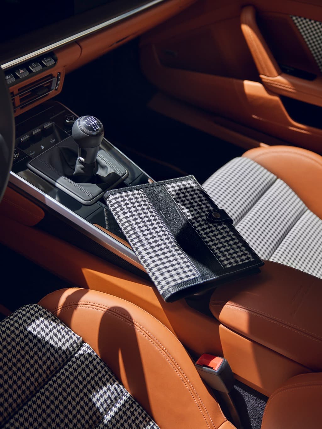 Black-and-white houndstooth notebook with a black leather strap on a brown Porsche leather seat next to the gear shifter