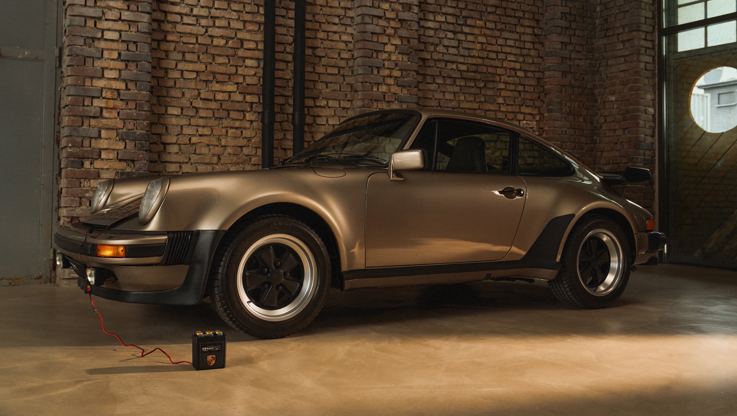 Classic gold Porsche 911 in a brick garage, connected to a battery charger on the floor.