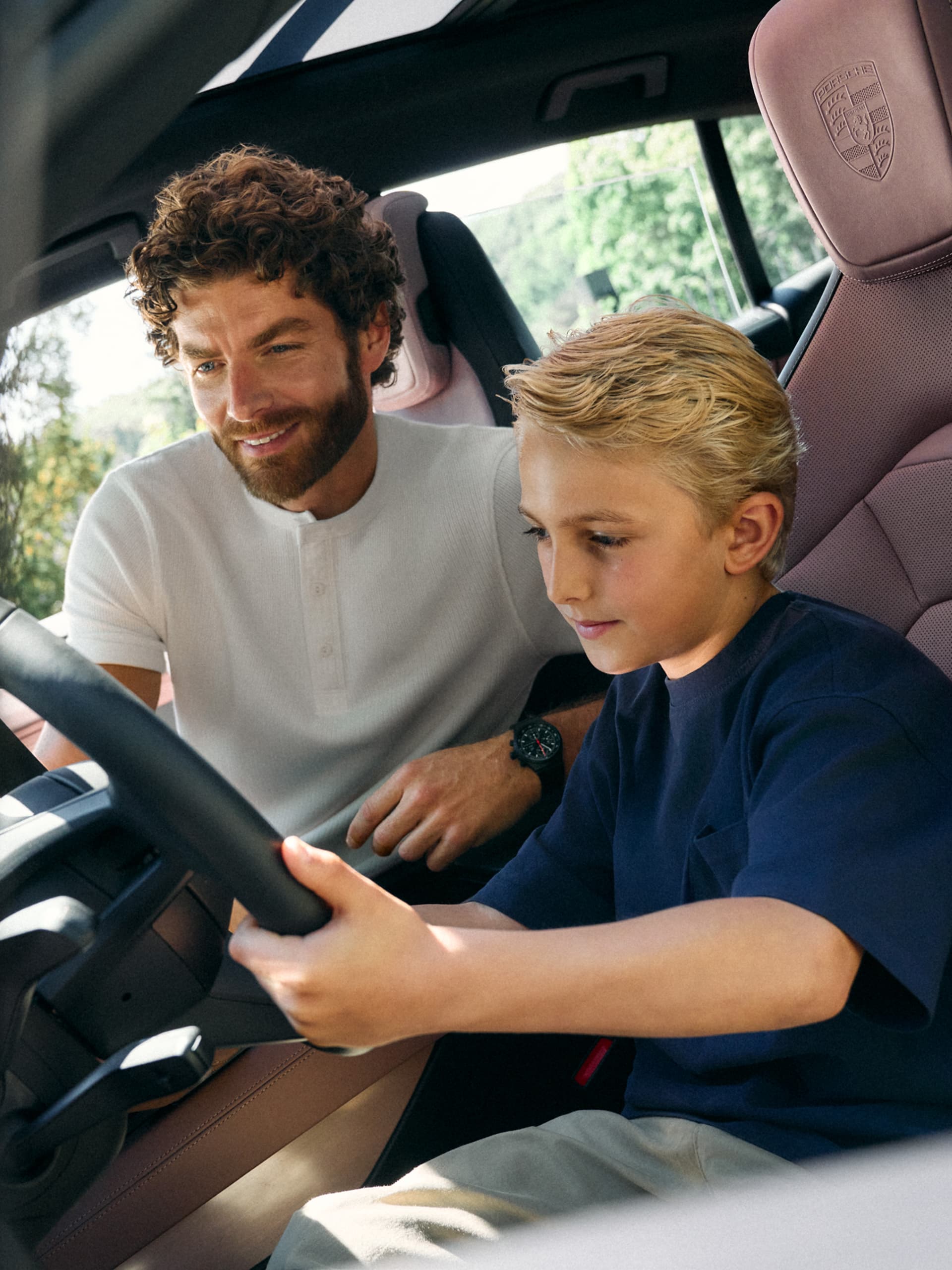 Padre y niño en un Porsche; el niño sujeta el volante; asientos de cuero rosa con el escudo de Porsche.