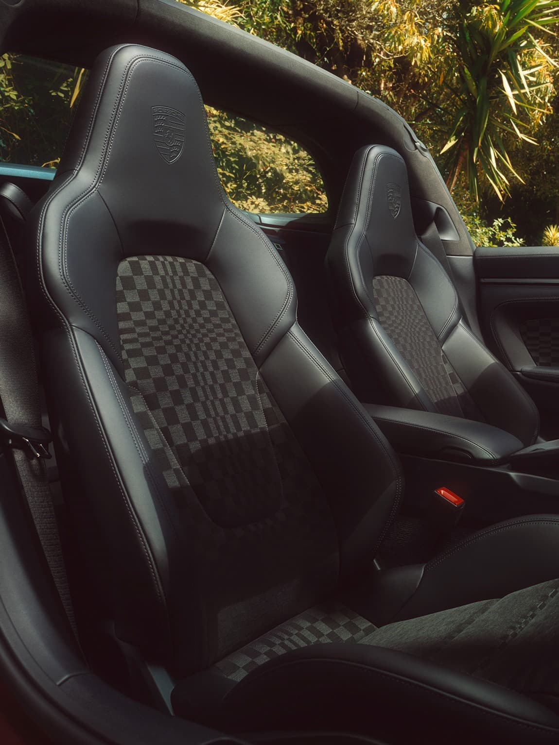 Black Porsche sports seats with chequered fabric inserts and an embossed crest; a convertible interior set against a backdrop of green plants.