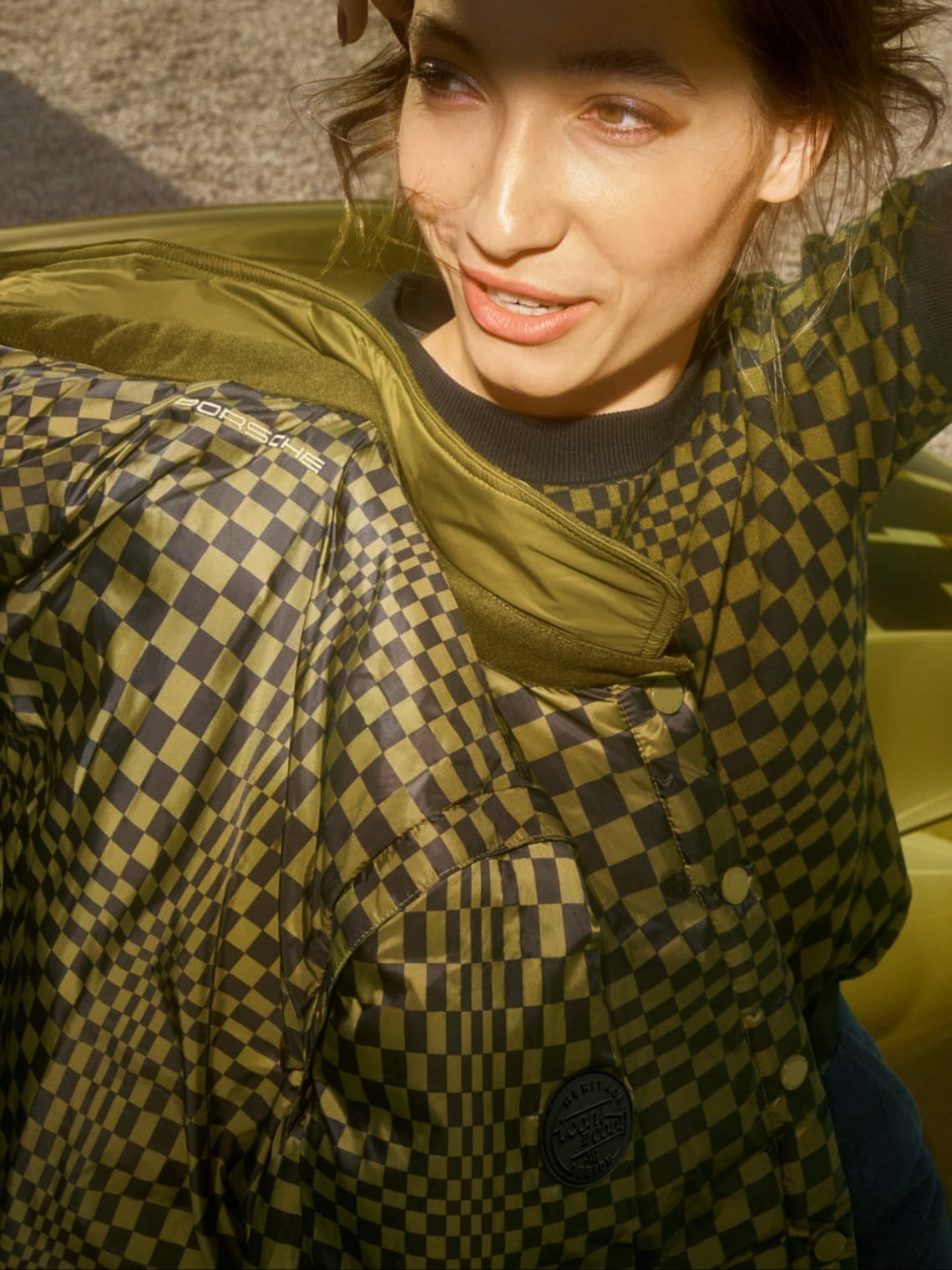 Model wearing olive and black checkered Porsche jacket with green lining, leaning on a car in sunlight.