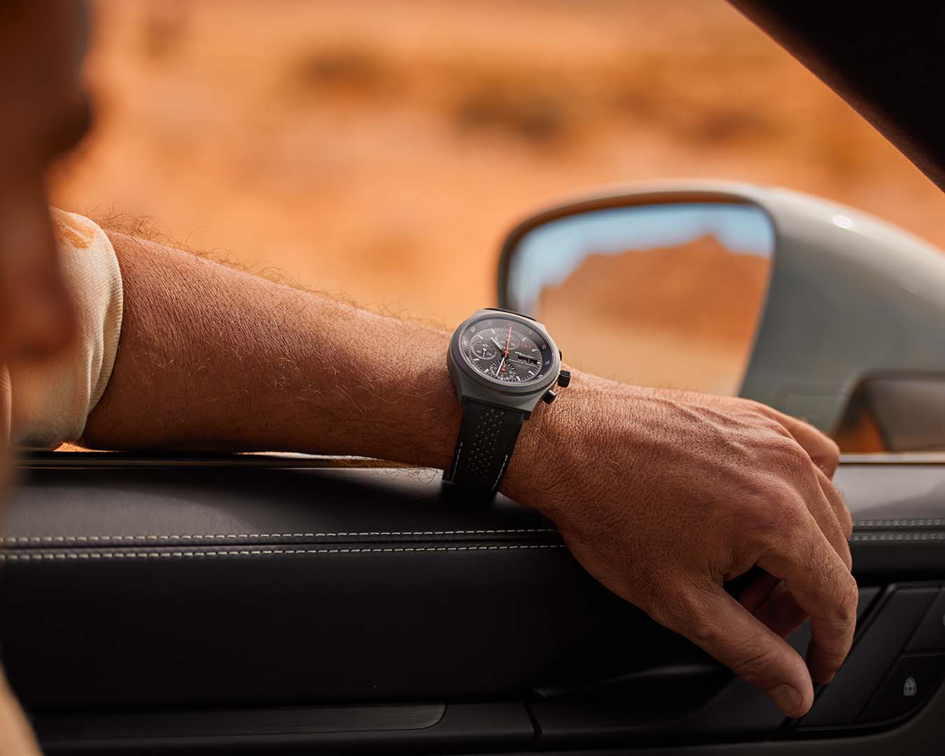 Mężczyzna siedzi w Porsche, opiera ramię o otwarte okno i ma na nadgarstku czarny zegarek Porsche 911 Dakar Chronograph; patrzy w dal.