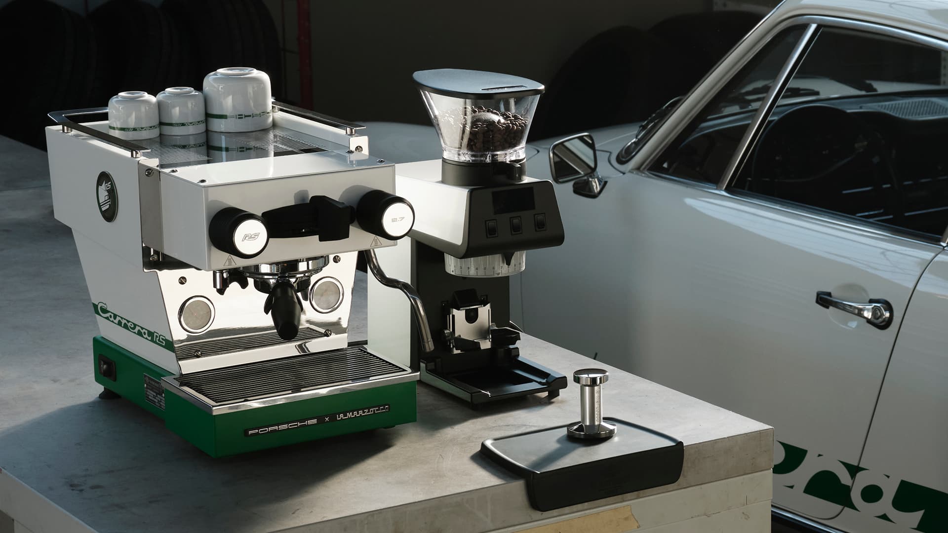 Machine à espresso La Marzocco blanche avec accents verts, moulin à café et tamper sur un établi, à côté d’une Porsche argentée.