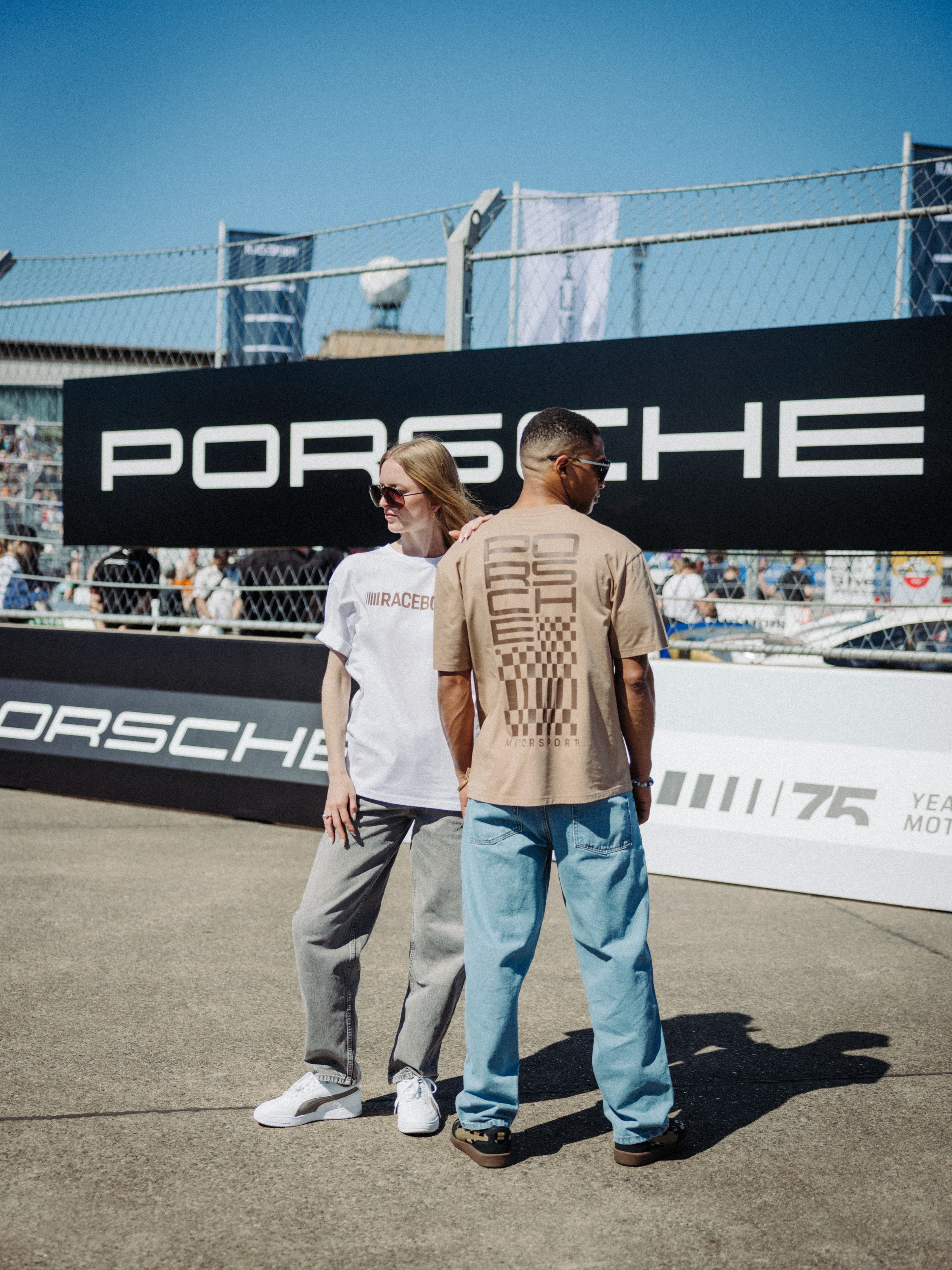 Deux modèles portant des vêtements Porsche Motorsport lors d’un événement de sport automobile en plein air, avec une signalétique Porsche en arrière-plan.