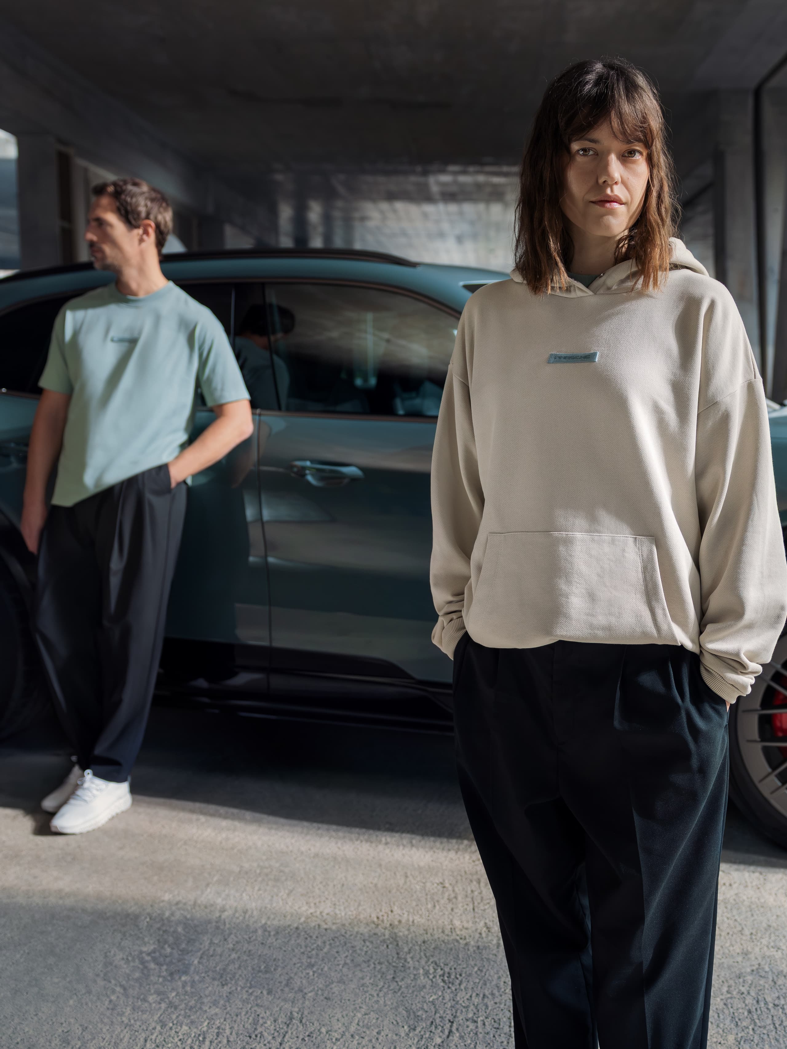 Two people pose casually in a concrete parking structure beside a blue‑green sporty SUV, creating a modern, urban lifestyle mood