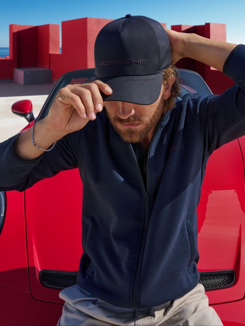Un homme est assis sur le capot d’une 911 S/C rouge, tenant sa casquette de la collection 911 S/C.