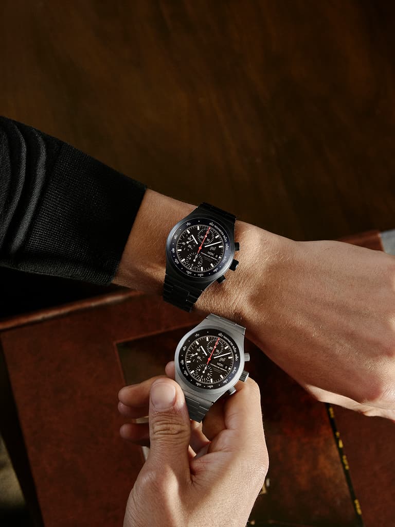 Due orologi da polso cronografi con quadrante nero, uno indossato al polso e uno tenuto in mano, su un tavolo in legno.