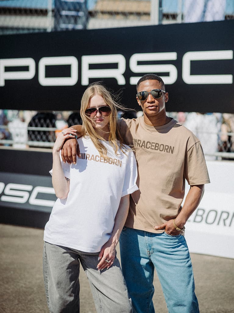 Due modelli con occhiali da sole che indossano T-shirt “RACEBORN” bianche e beige davanti a un banner Porsche all’aperto.