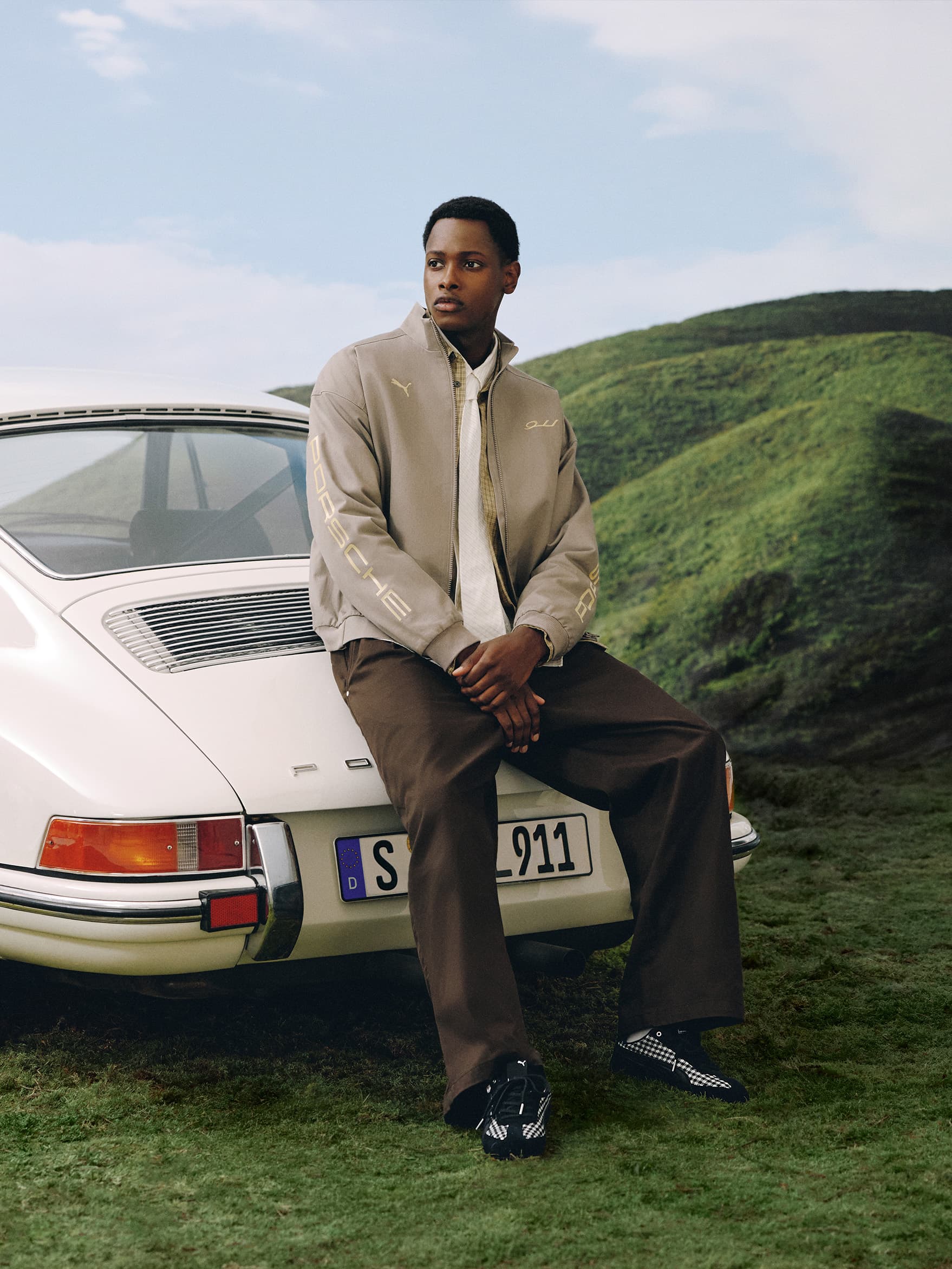 Model in beige Porsche jacket and brown pants sitting on white Porsche 911, green hills in background